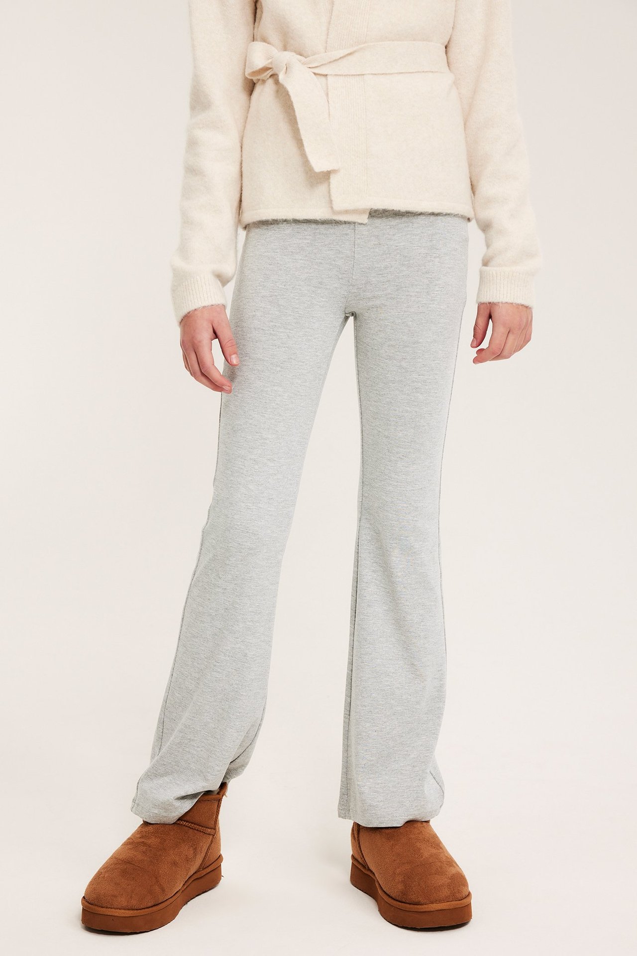 Etunäkymä: Lapsi yllään vaaleanharmaat meleeratut flare-leggingsit, kermanvärinen solmittava neuletakki ja ruskeat nilkkurit.