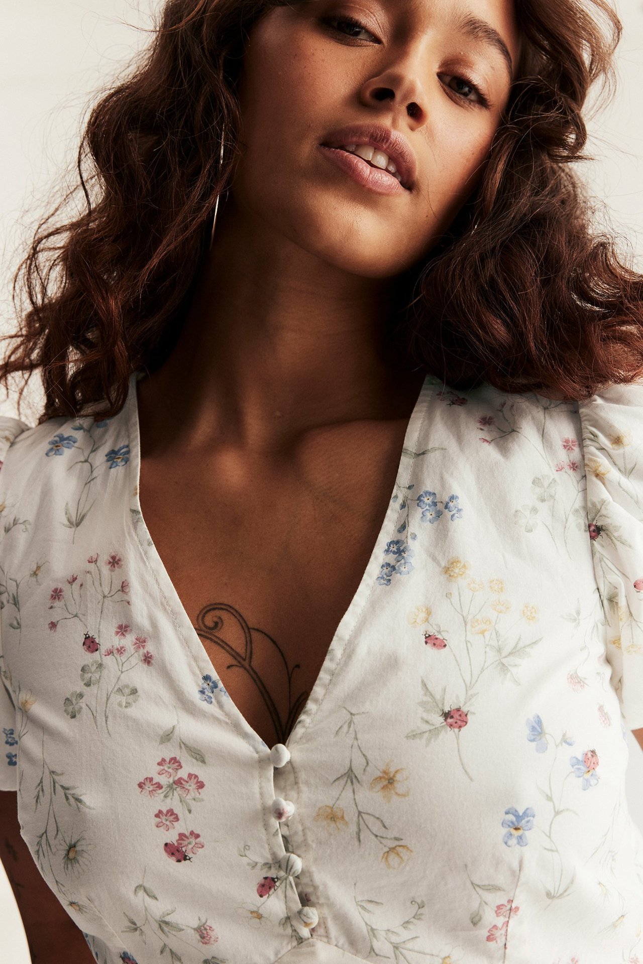 Nainen luonnonvalkoisessa kukkamekossa, V-kaula-aukko, päällystetyt napit ja pussihihat, yllään renkaat korvissa.