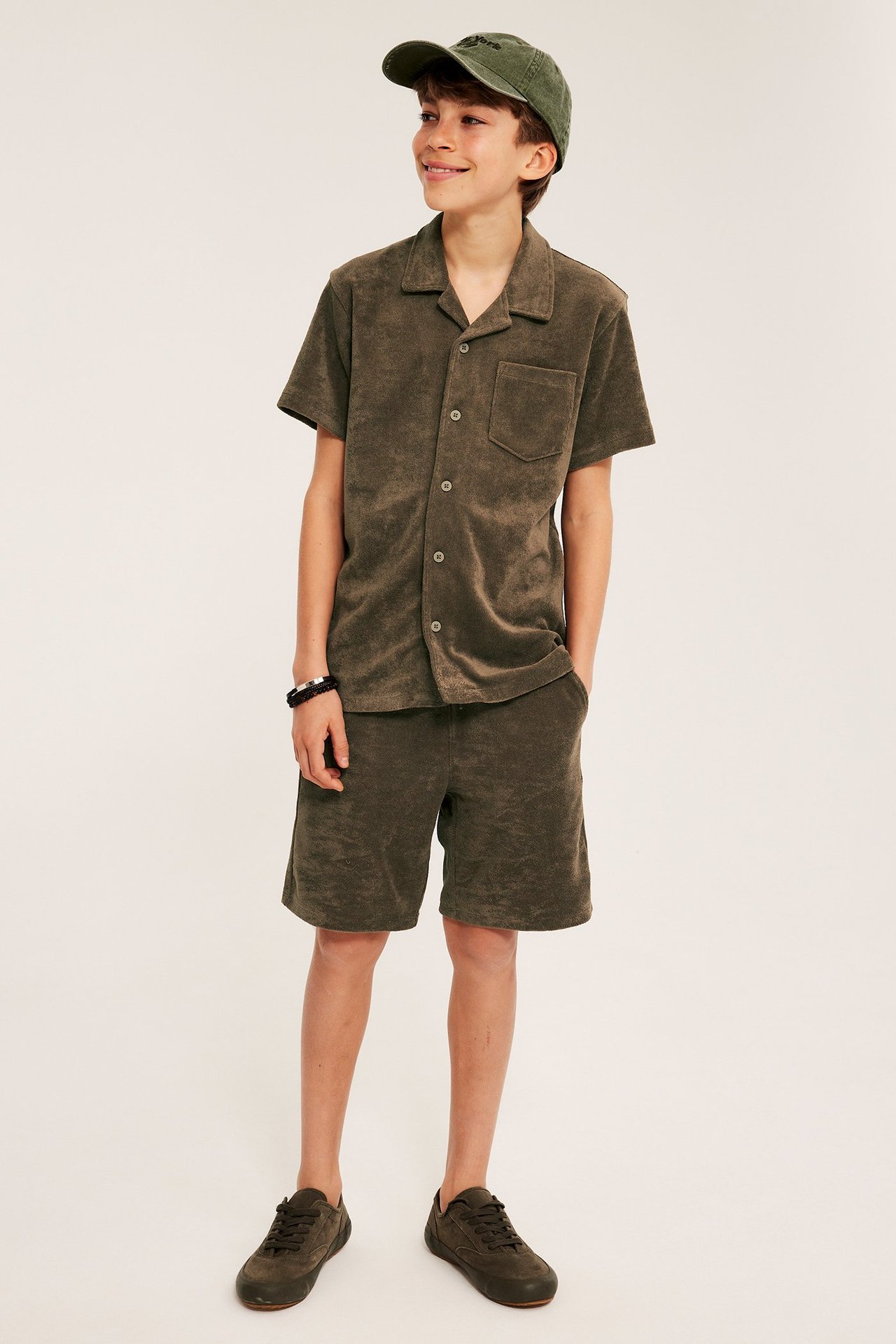 Barn, framifrån: brun frottéskjorta, matchande shorts, grön keps och sneakers.