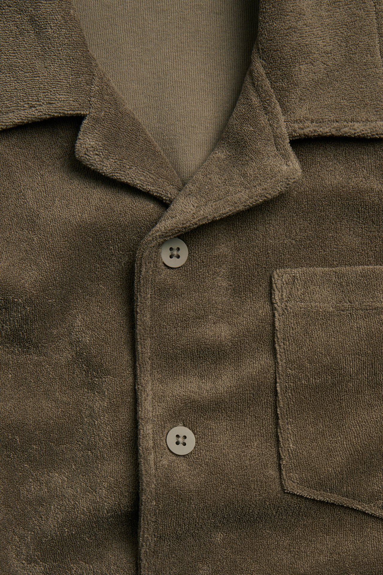 Närbild: Brun strukturerad frottéskjorta med resortkrage, bröstficka och knappar, över en ljusare t-shirt.