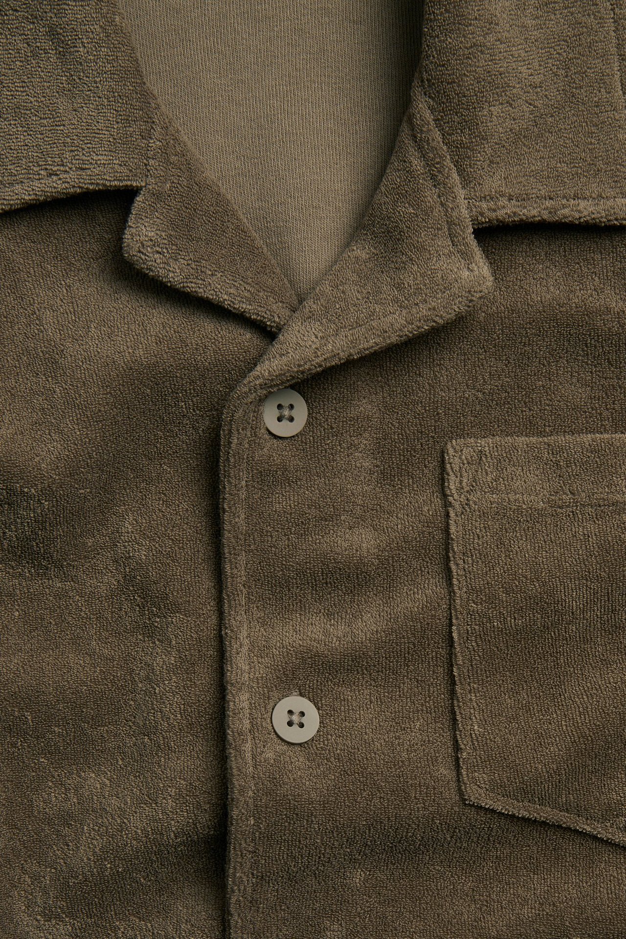 Lähikuva: Ruskea froteepaita resort-kauluksella, rintataskulla ja napeilla, vaaleamman t-paidan päällä.