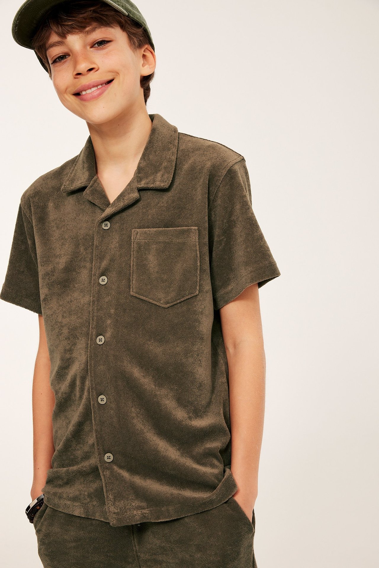 Frontvy: Barn bär brun kortärmad frottéskjorta, resortkrage, bröstficka, matchande shorts, grön keps.