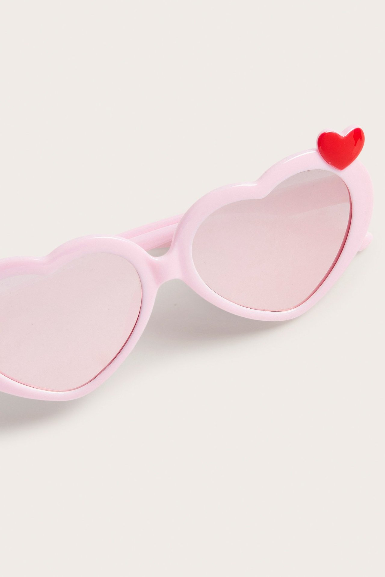 Lähikuva vaaleanpunaisista muovisista sydämenmuotoisista aurinkolaseista ja punaisesta sydändetaljista.