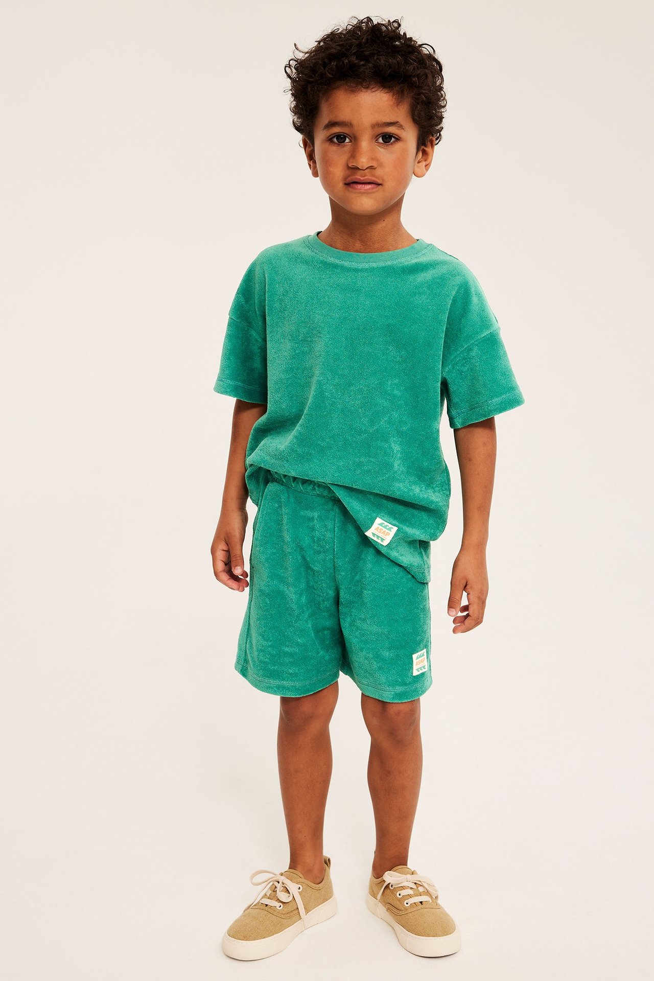Lapsi edestä, vihreät froteeshortsit, niihin sopiva t-paita ja beiget lenkkarit.