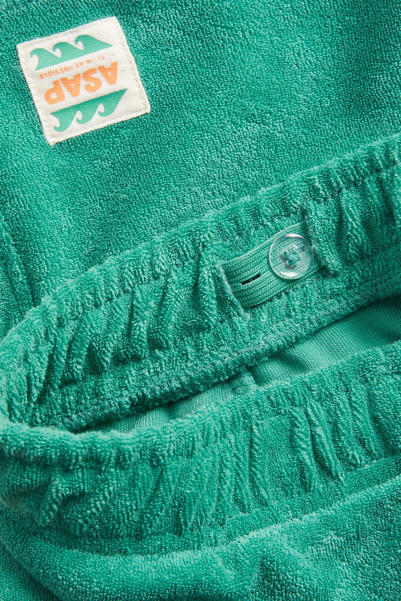 Lähikuva vihreistä froteeshortseista, joissa joustava vyötärö, säädettävä nappi ja kudottu etiketti.