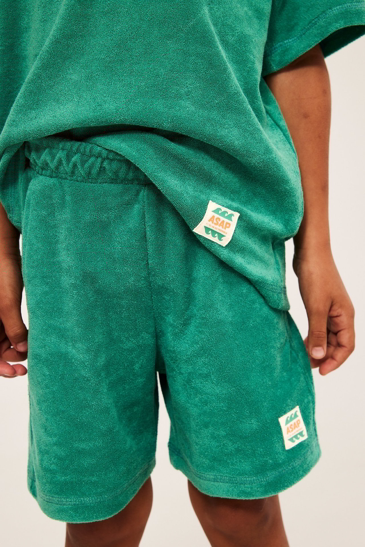 Lähikuva edestä lapsesta yllään löysät vihreät froteeshortsit sivutaskuilla ja yhteensopiva t-paita.