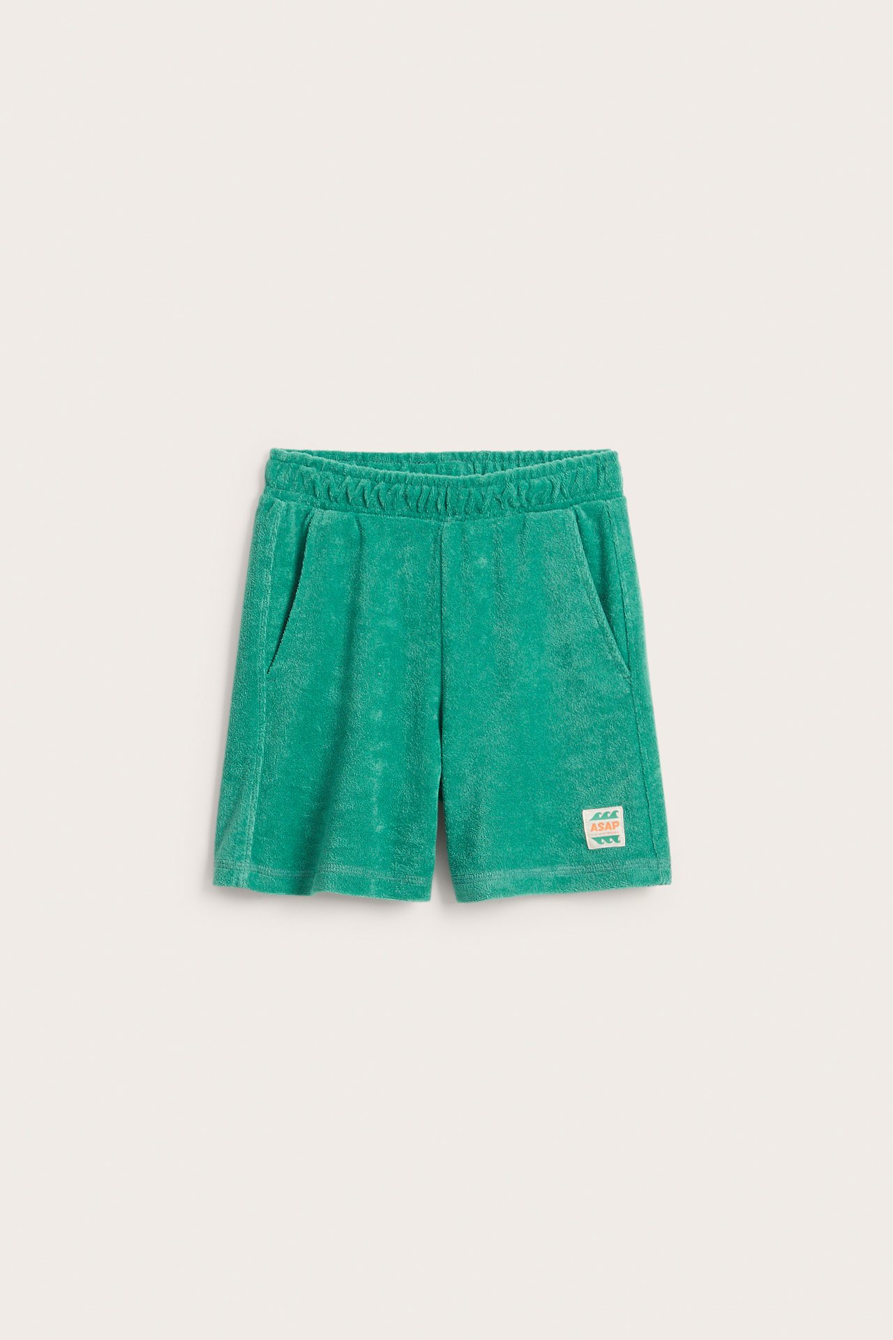 Edestäpäin: vihreät froteeshortsit lapselle, joissa joustava vyötärö, sivutaskut ja pieni värikäs etiketti.