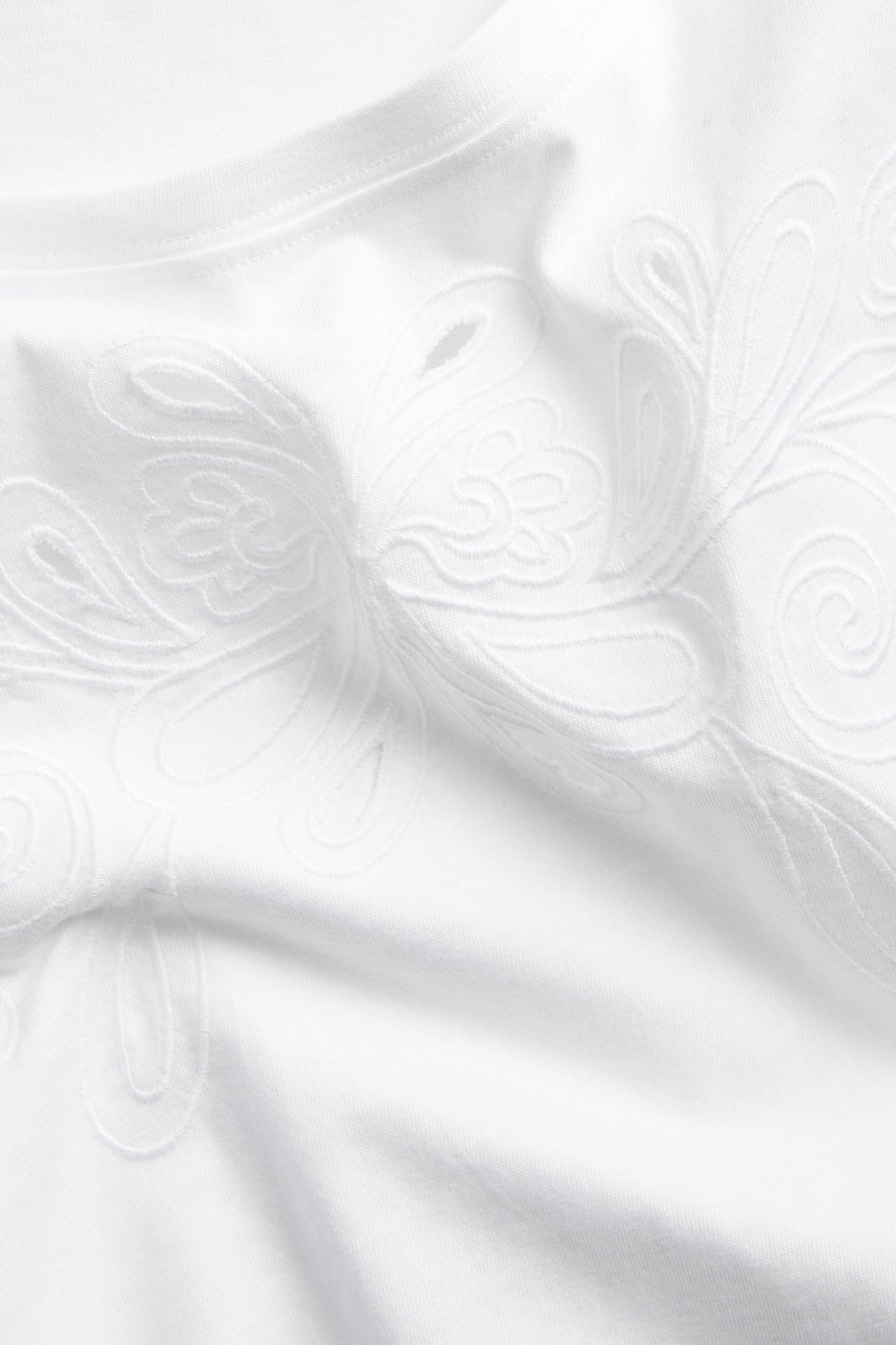 Lähikuva valkoisesta puuvilla-T-paidasta, jossa on yksityiskohtainen broderie anglaise -kirjailu.