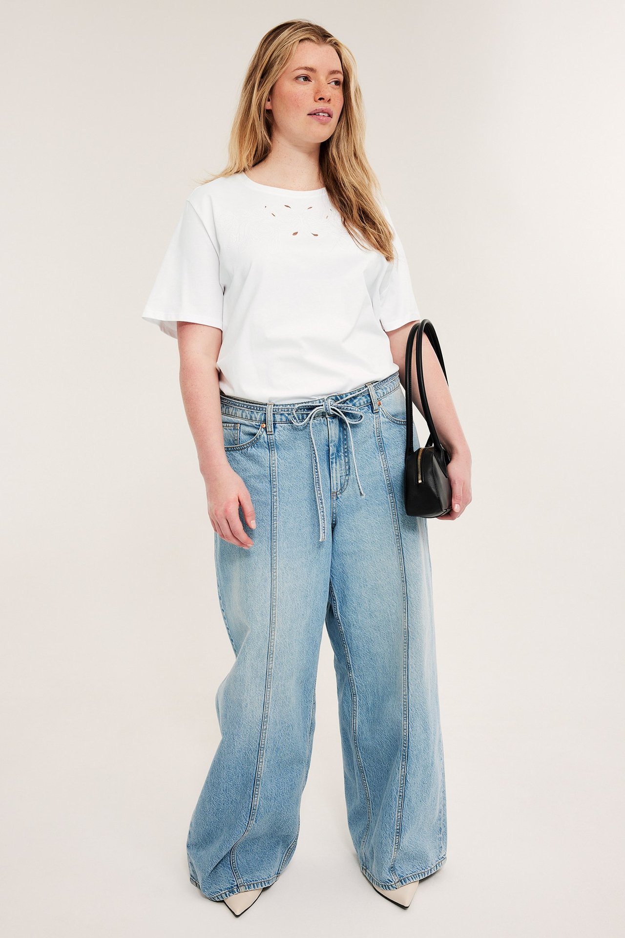 Framsida: kvinna i vit broderie anglaise T-shirt, ljusblå vida jeans, håller svart handväska.