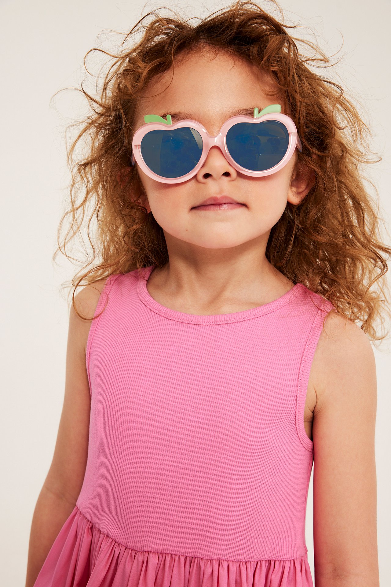 Lähikuva edestä: Lapsi yllään vaaleanpunaiset omenanmuotoiset aurinkolasit vihreillä yksityiskohdilla ja pinkki ribbitoppi.