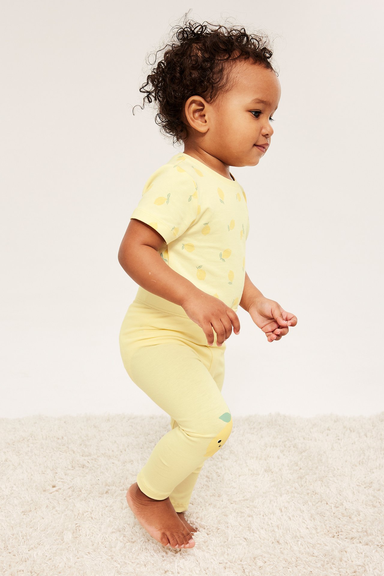 Widok z boku: niemowlę w jasnożółtych legginsach z aplikacjami na kolanach i pasującym body w cytrynowy wzór.