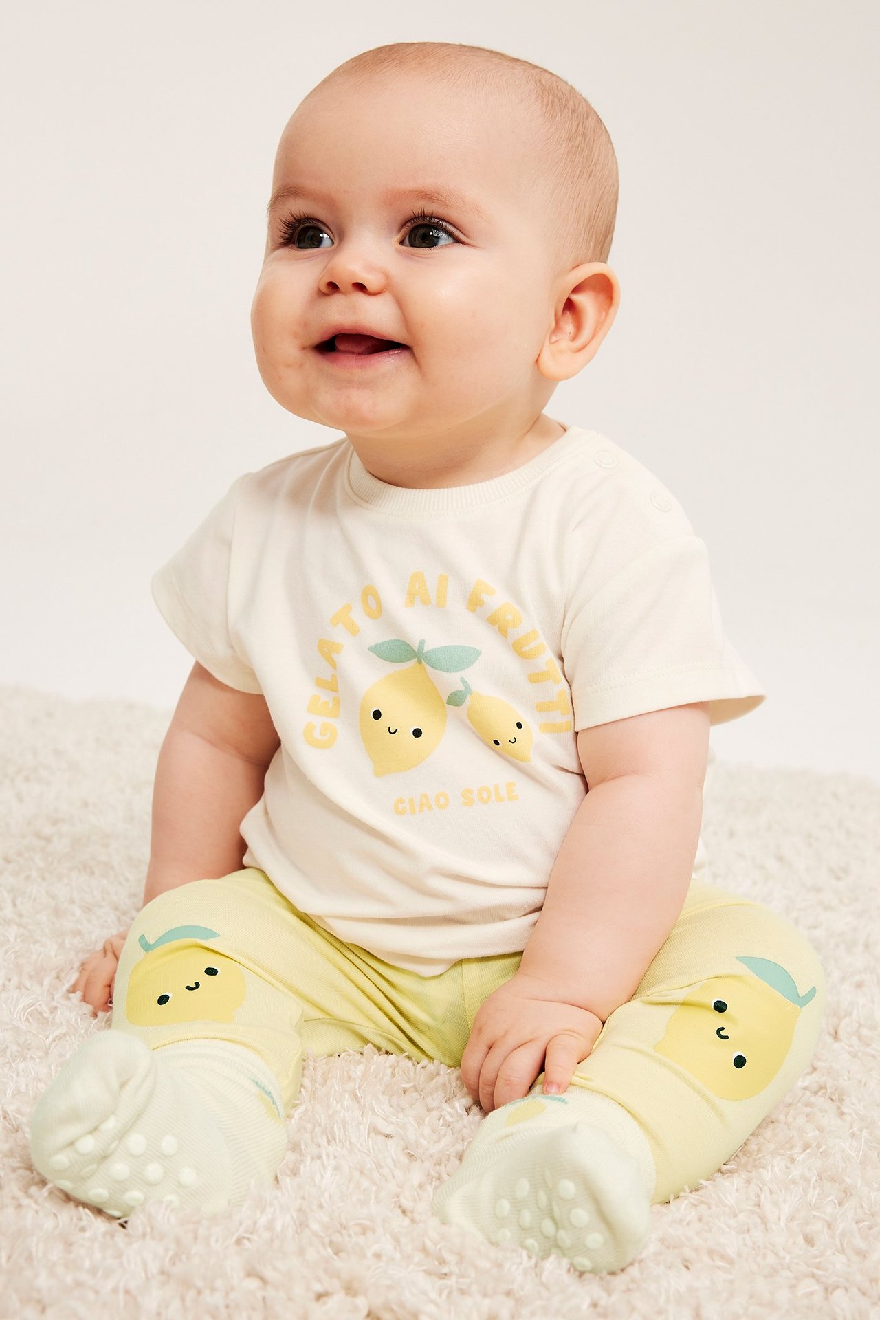 Widok z przodu: niemowlę w jasnożółtych legginsach z cytrynami, beżowej koszulce i zielonych skarpetkach antypoślizgowych.