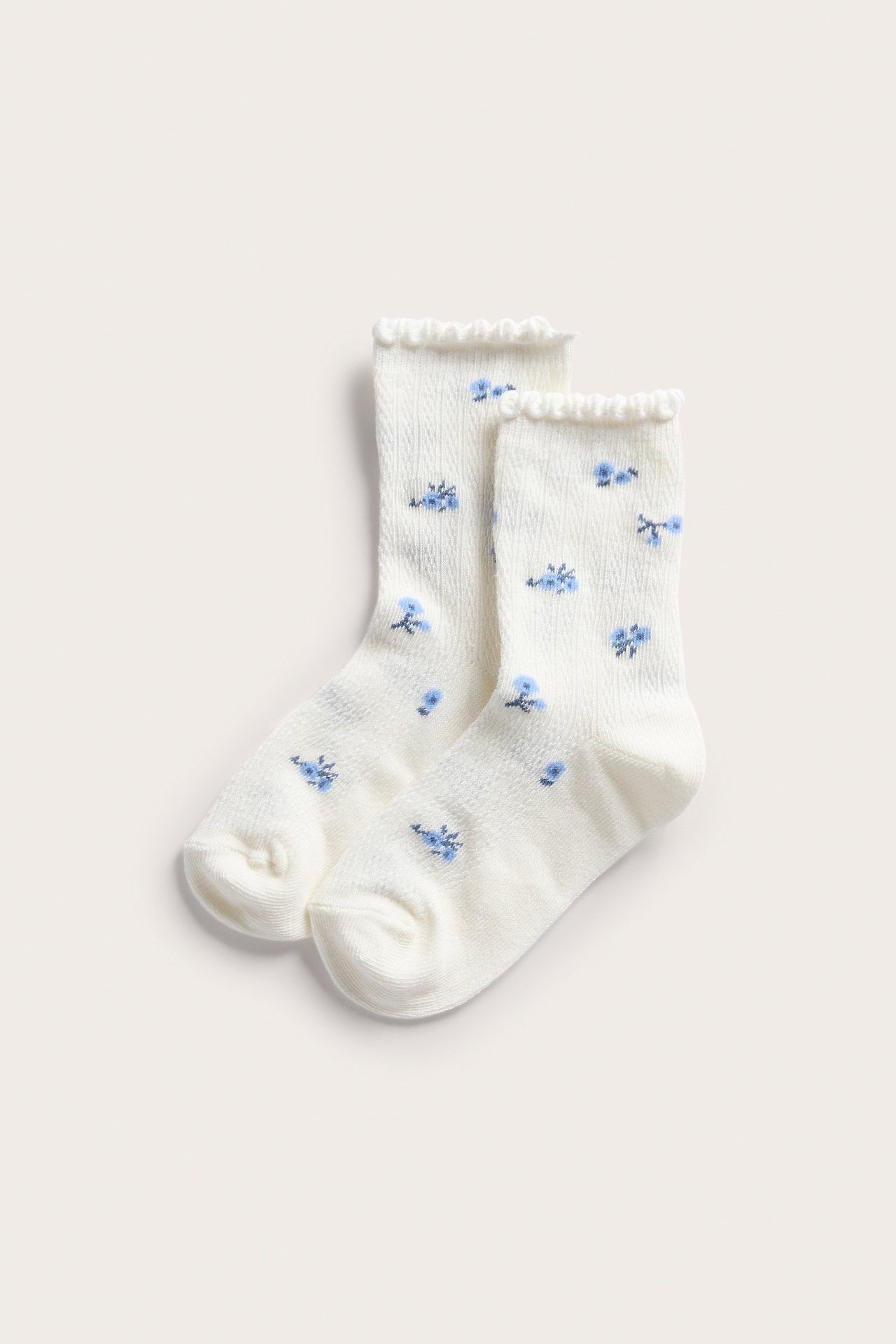 Etunäkymä kahdesta luonnonvalkoisesta lasten sukasta, joissa sininen kukkakuvio ja röyhelöreunat.