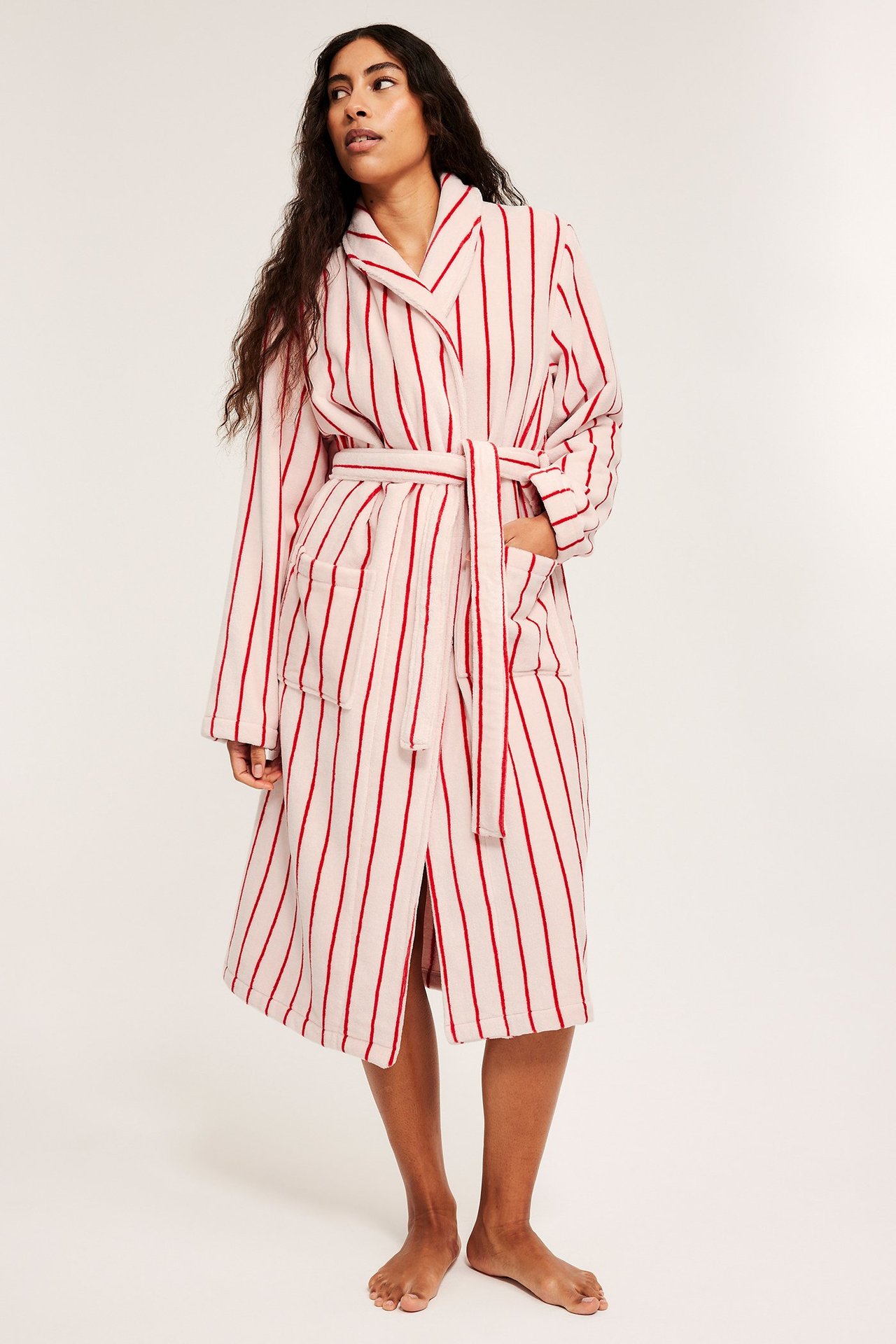 Framifrån, kvinna i ljusrosa bomullsfrottémorgonrock med röda ränder och knytskärp.