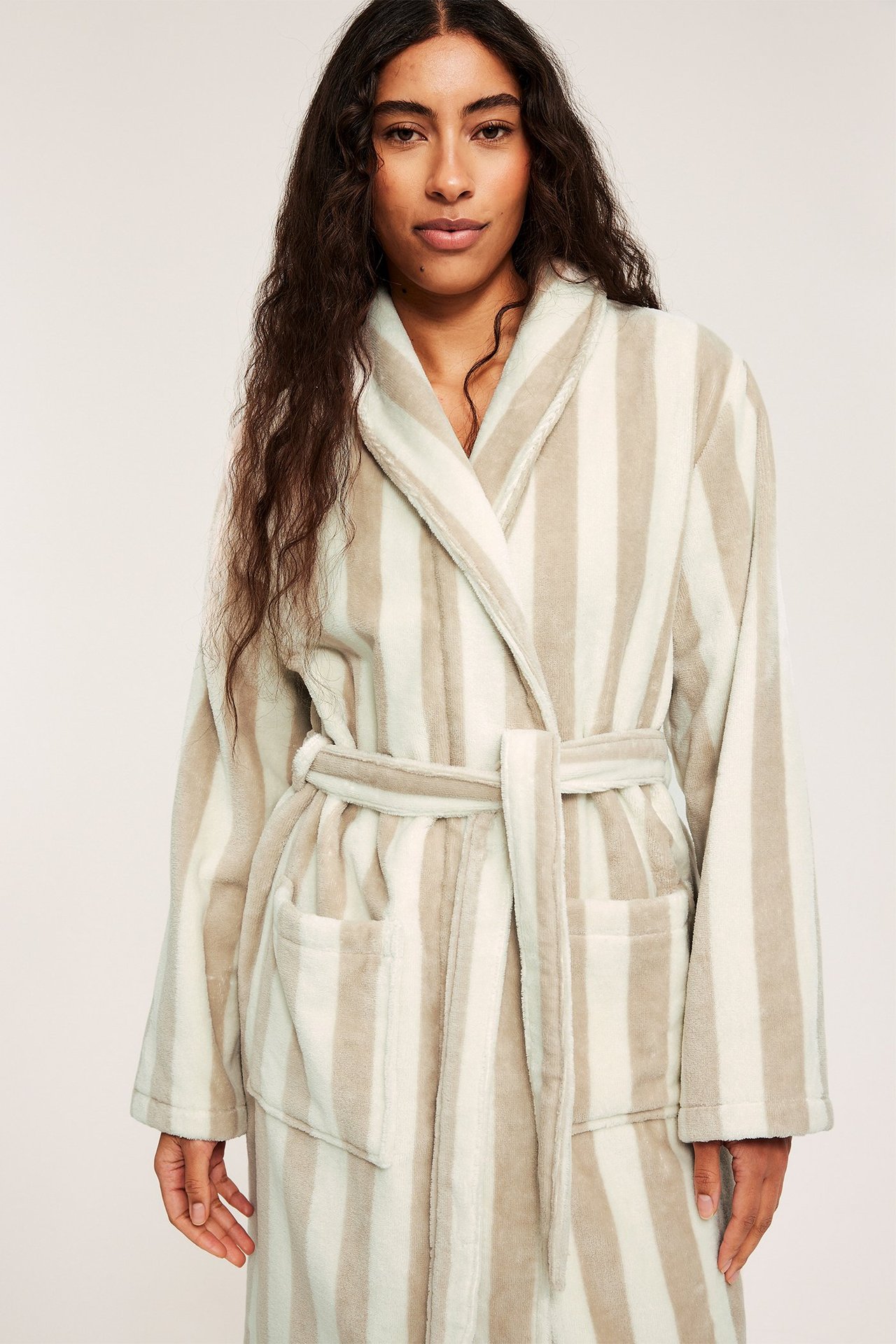 Pehmeä beige-valkoraidallinen froteeaamutakki, jossa naisella on huivikaulus, taskut ja vyö.