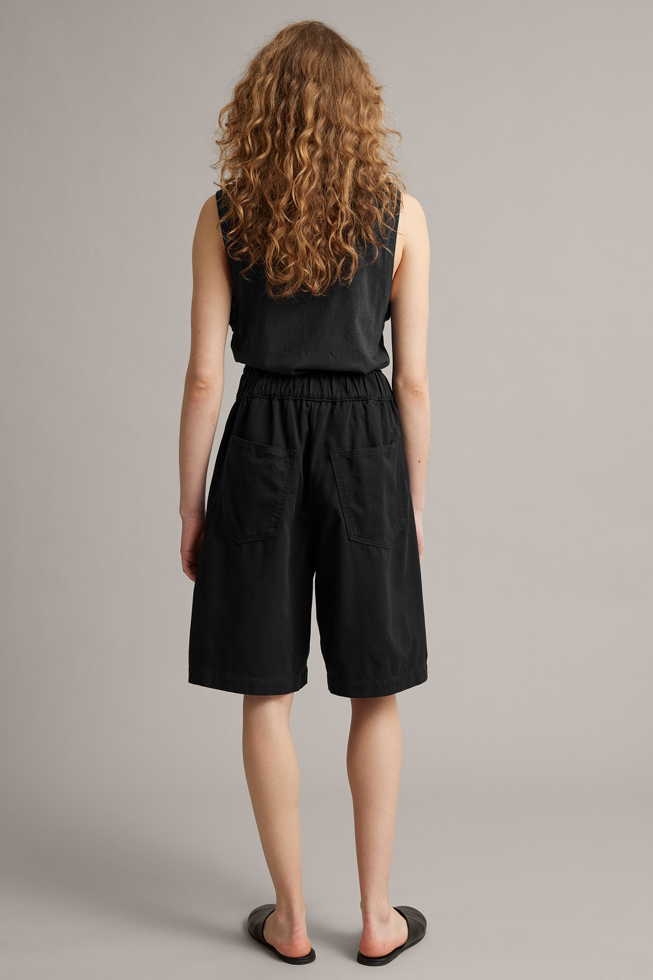 Kvinna sedd bakifrån i svarta bermudashorts, svart ärmlös topp och svarta platta sandaler.