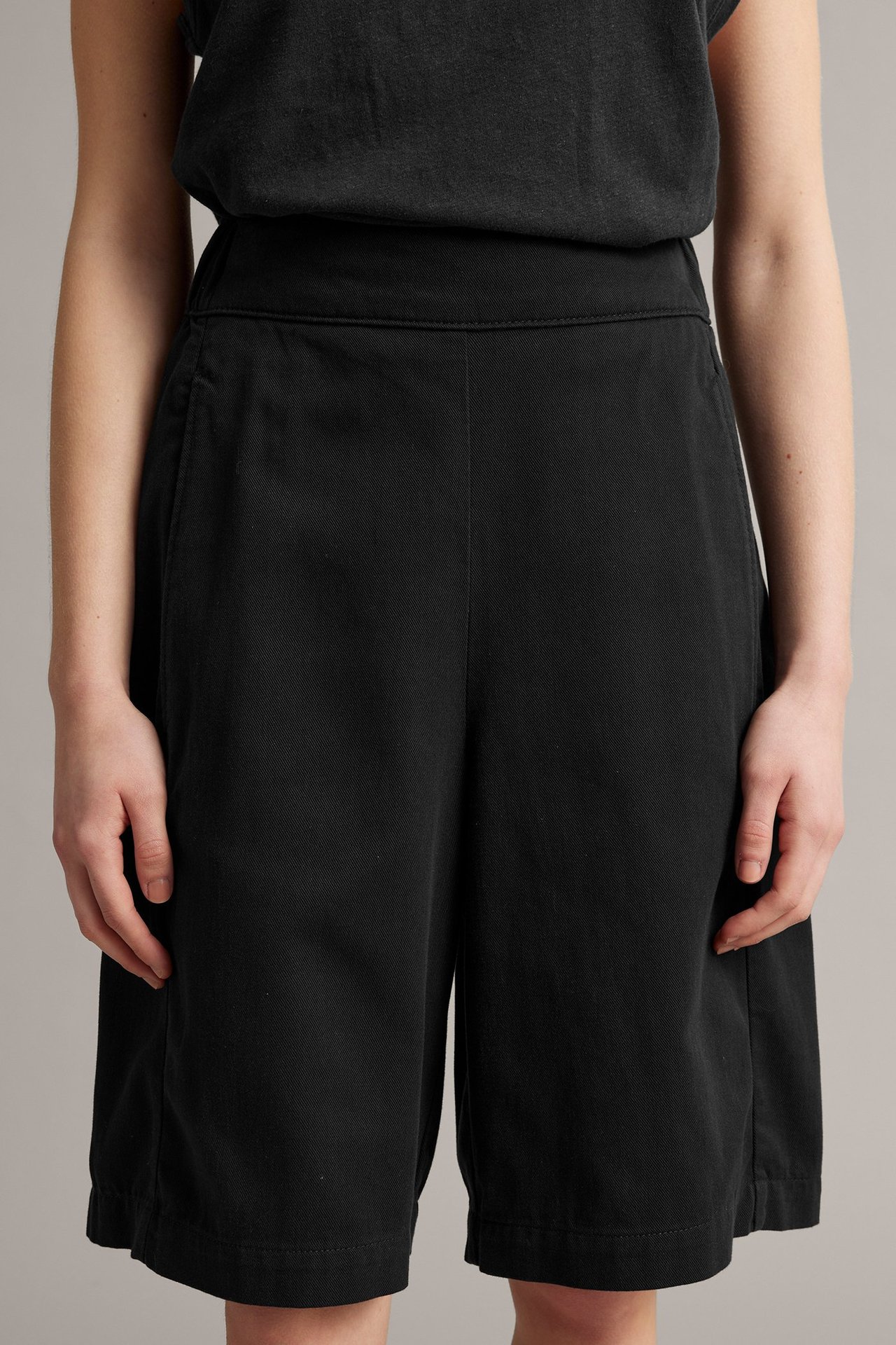 Framifrån, kvinna bär svarta lediga bermudashorts med hög midja och en svart topp.