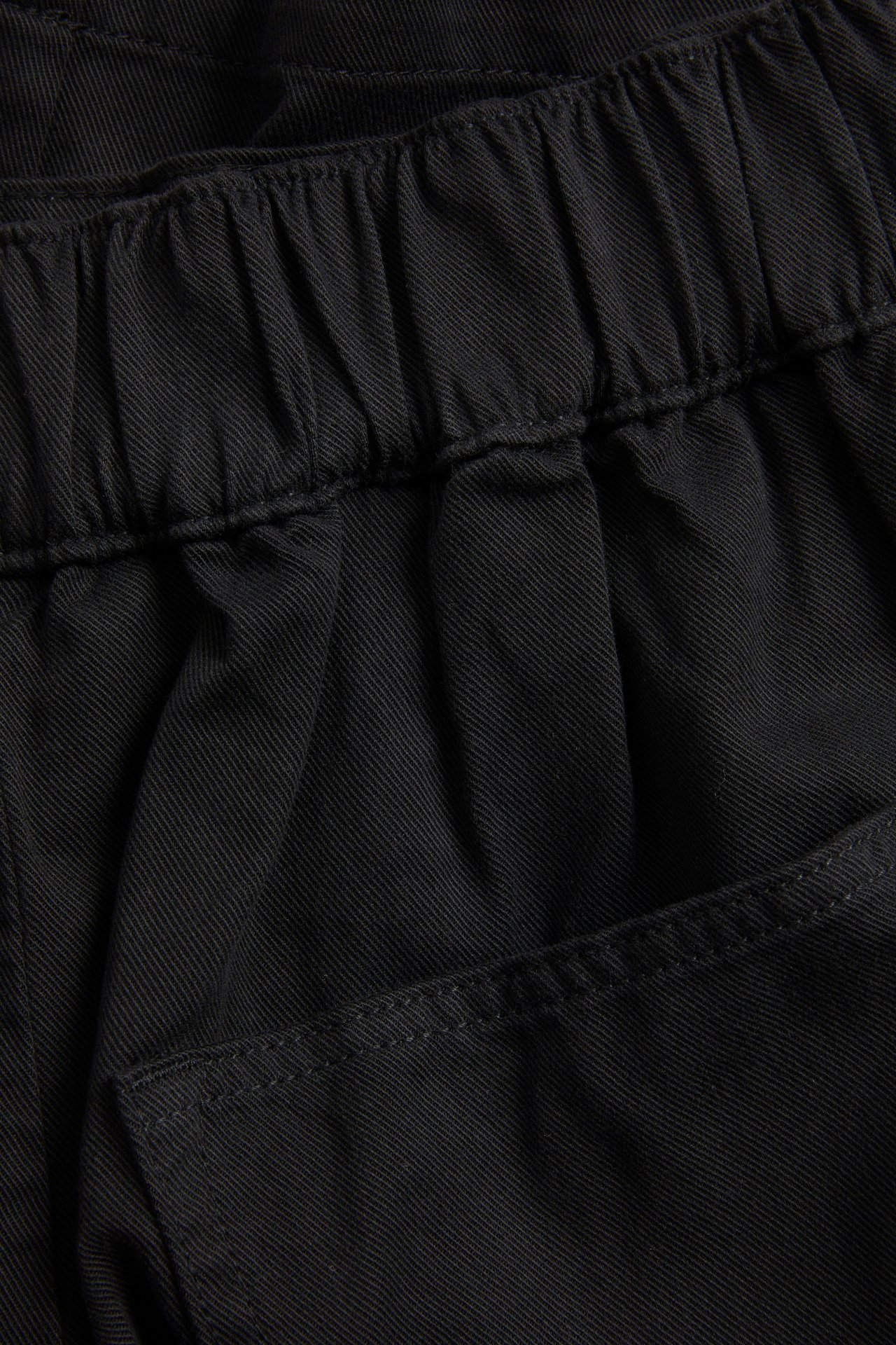 Lähikuva yksivärisistä mustista lyocell- ja puuvillabermuda-shortseista, joissa joustovyötärö ja taskuyksityiskohta.