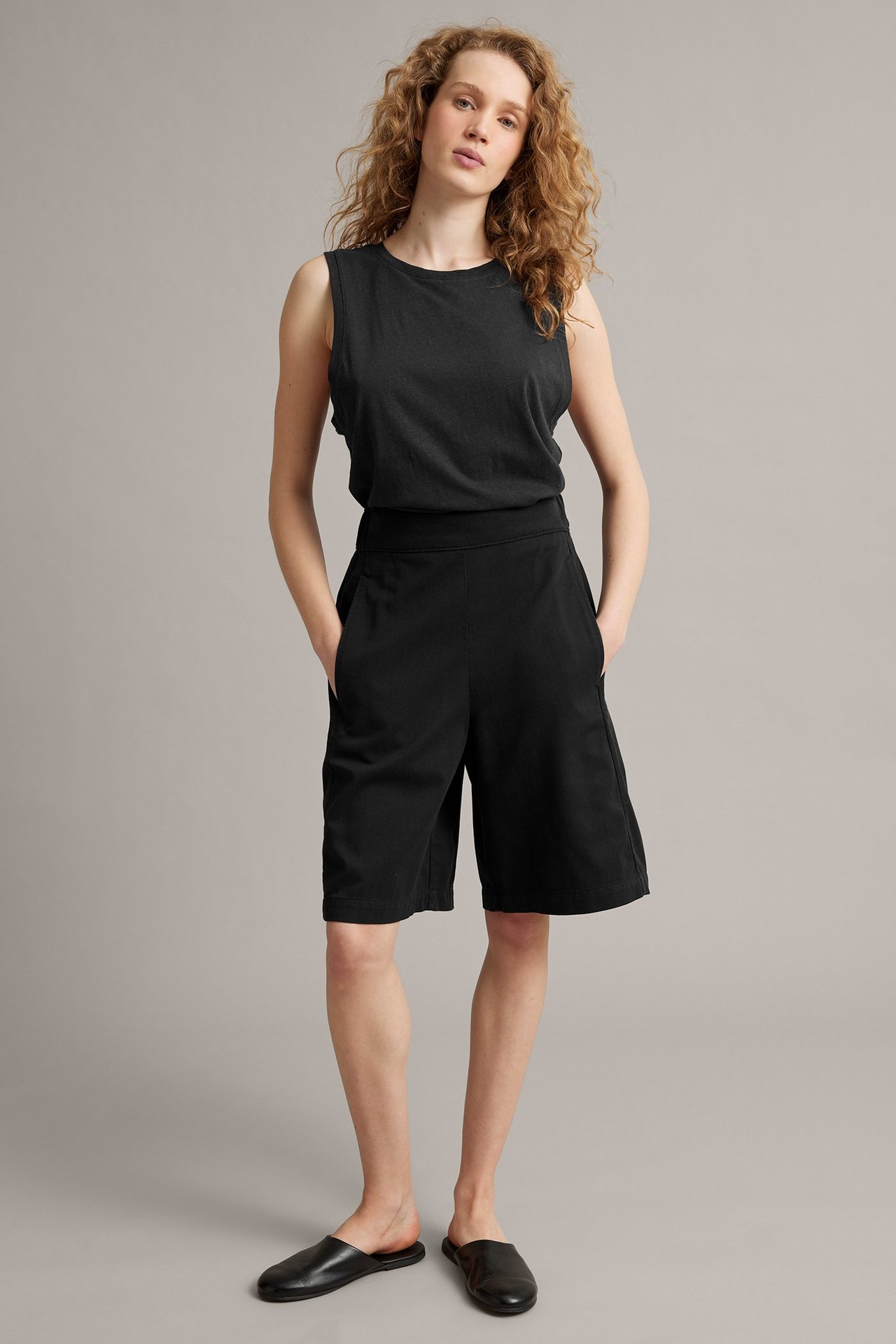 Pełny widok kobiety w czarnych szortach bermudach lyocell-bawełna, czarnym topie bez rękawów, czarnych klapkach.