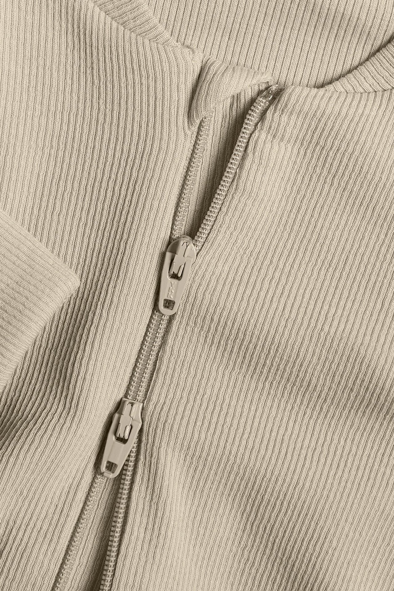 Lähikuva beigestä ribbipuuvillaisesta vauvanhaalarista kaksisuuntaisella vetoketjulla.