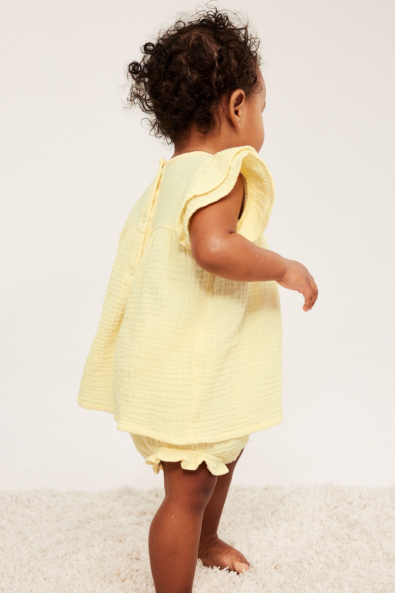 Sidovy: baby i ljusgult set i bomullsmuslin med topp med volangärmar, tryckknappar och matchande bloomers.