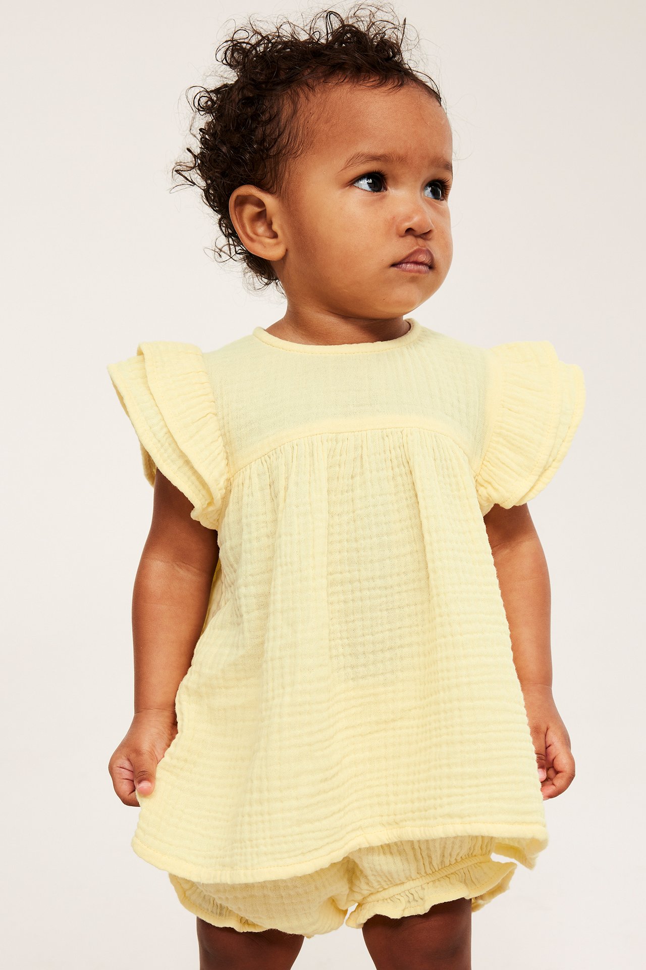 Framifrån, en baby i ljusgult set av bomullsmuslin, med topp med volangärmar och matchande bloomers.
