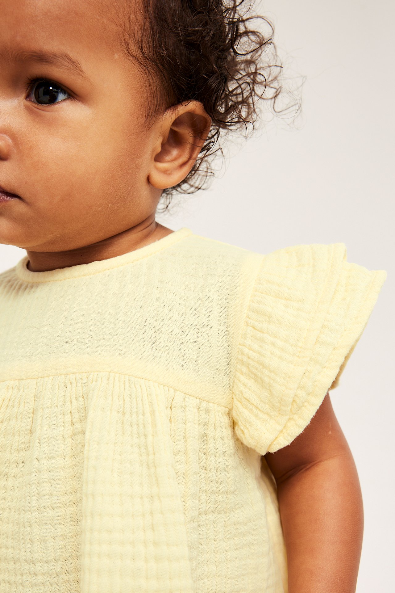 Nærbilde av en baby i lys gul topp av bomullsmuslin med rysjeermer og samlede detaljer.