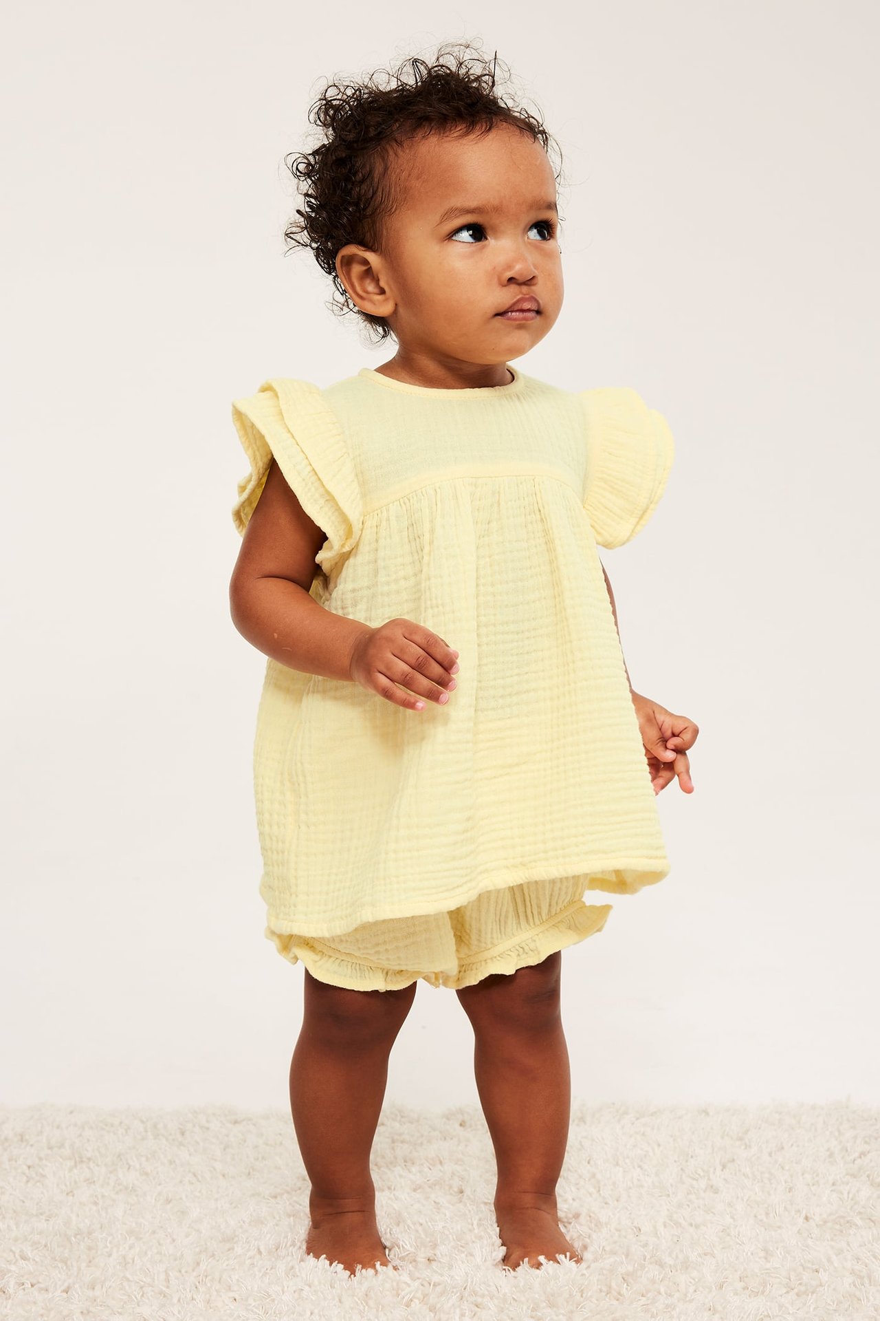 Framifrån, baby i ljust gult bomullsmuslinset med volangtopp och matchande bloomers.