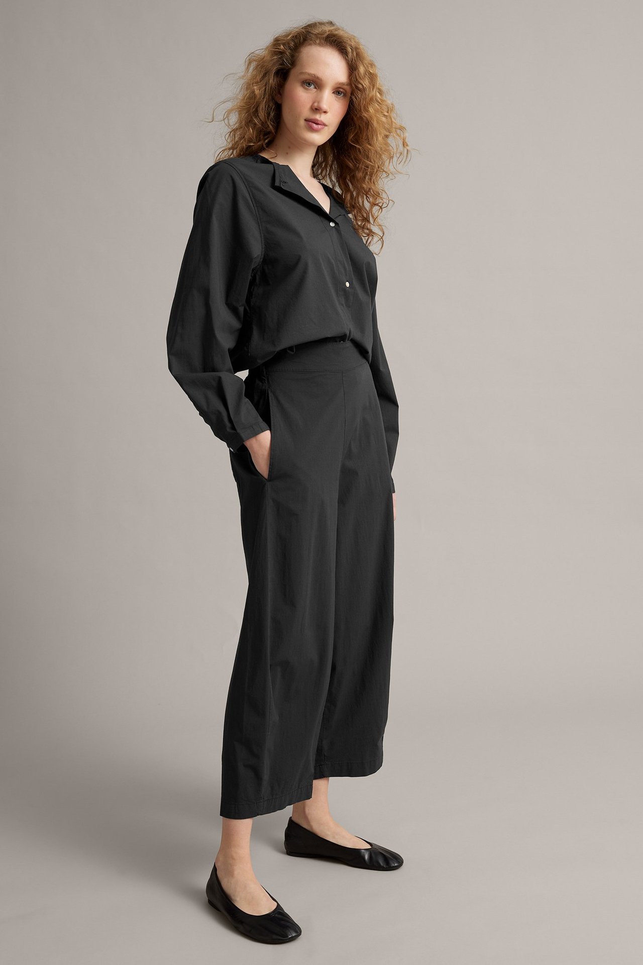 Sidovy av kvinna i svarta barrel-fit poplinbyxor, svart skjorta och svarta ballerinaskor.