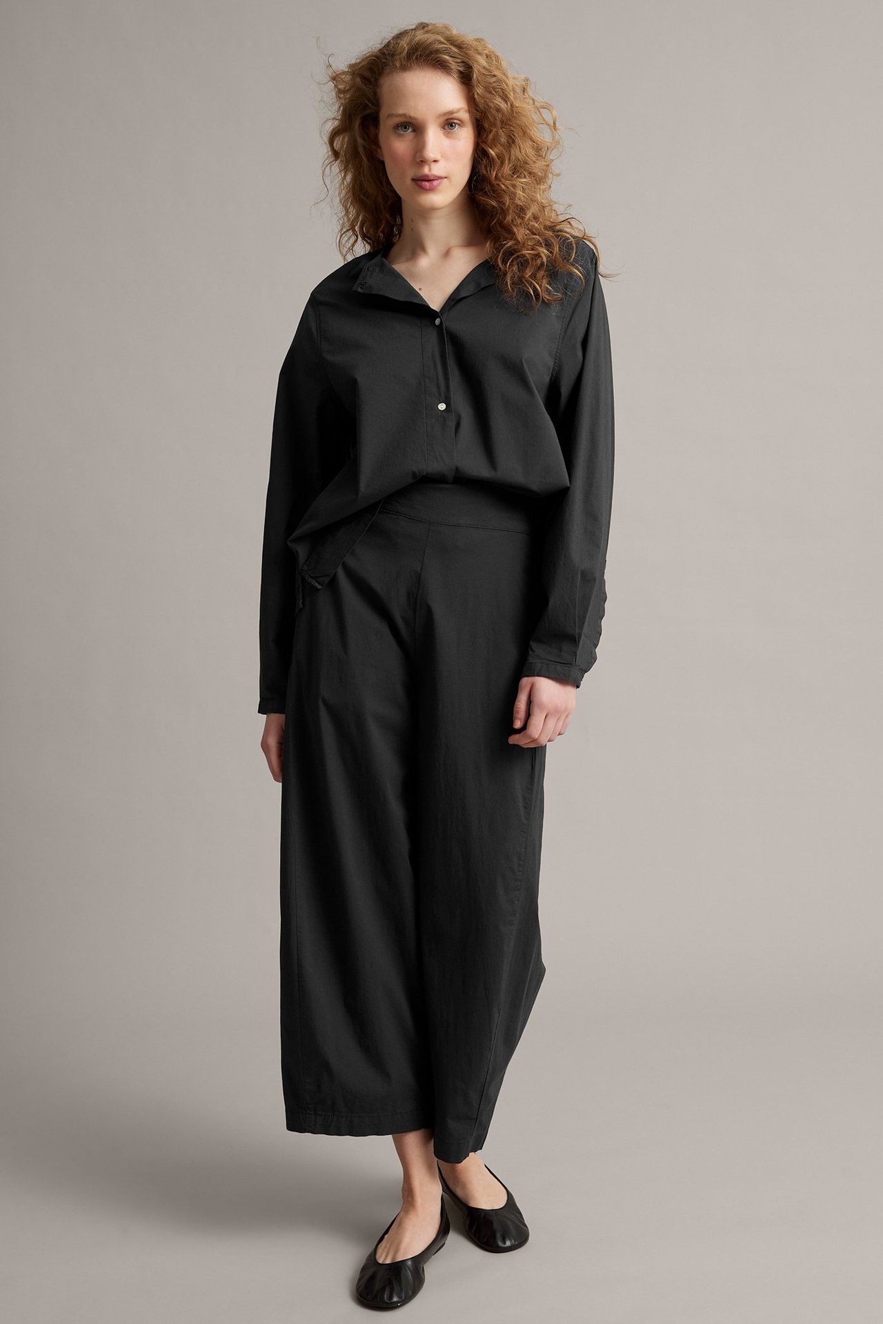 Widok z przodu: kobieta w czarnych spodniach barrel fit, czarnej koszuli i czarnych balerinkach.