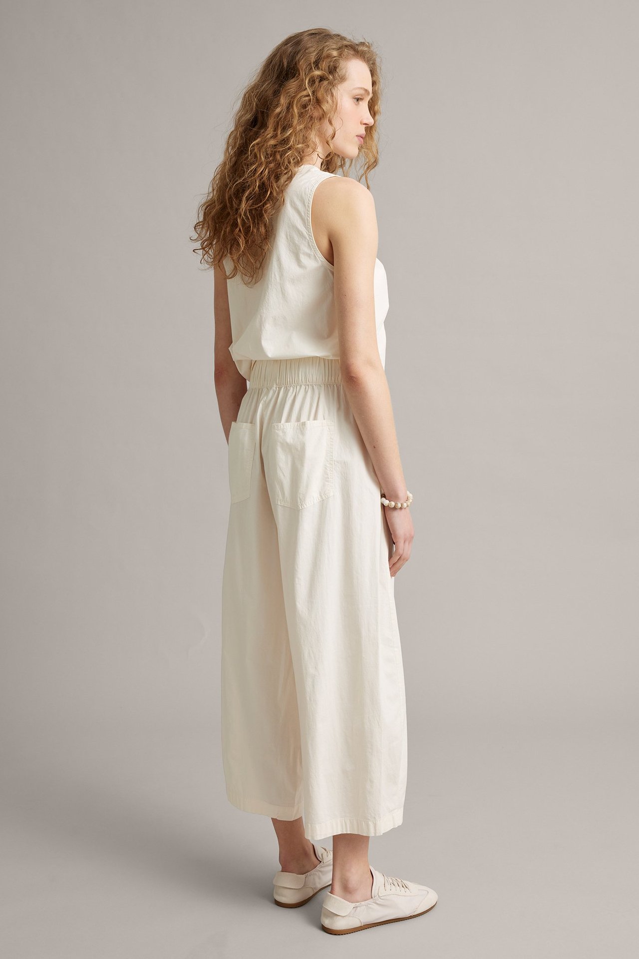 Sivukuva: Nainen valkoisissa barrel-fit housuissa joustovyötäröllä, yhteensopiva hihaton toppi ja valkoiset tennarit.