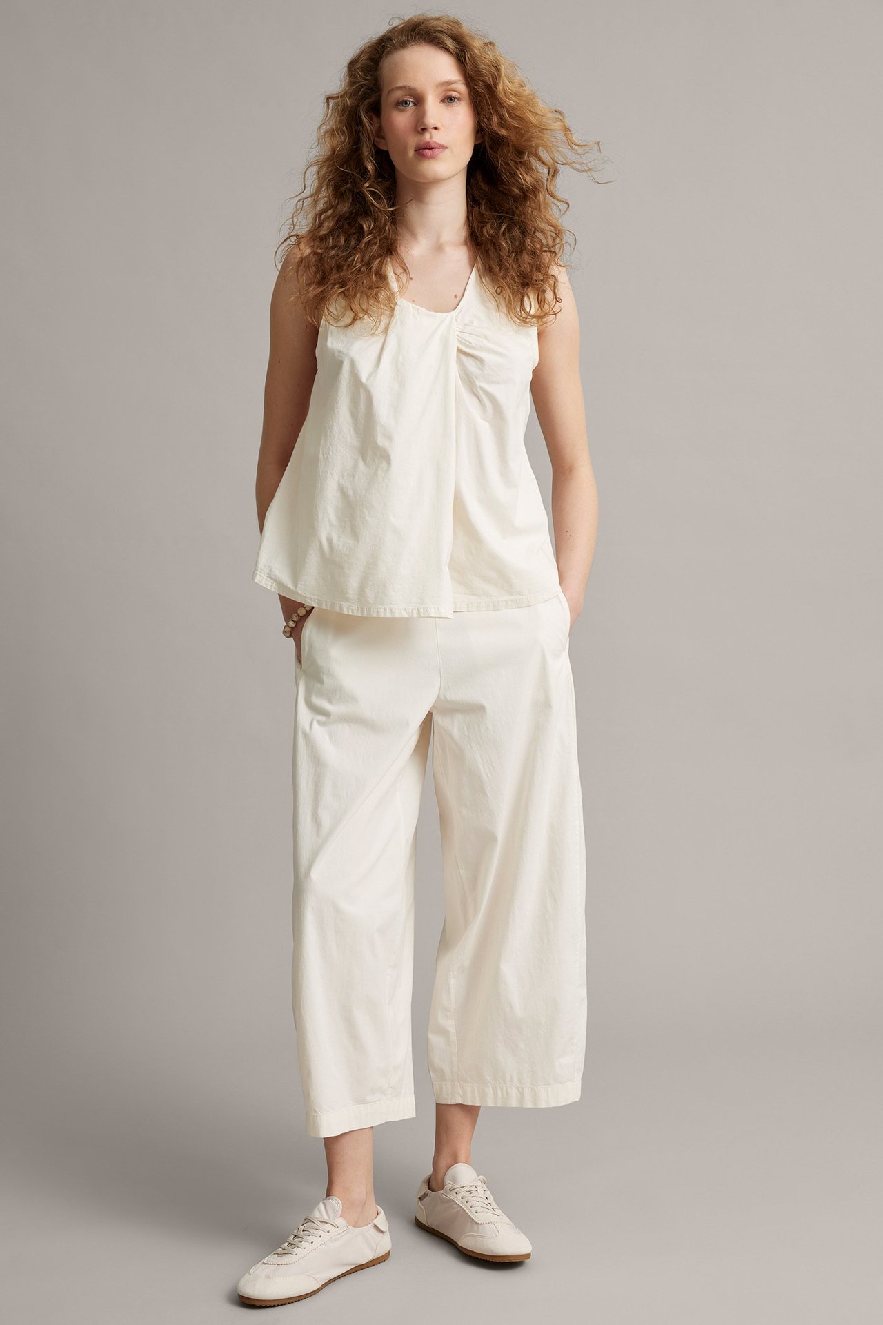Kobieta od przodu w białych spodniach barrel-fit z kieszeniami, dopasowanym topie bez rękawów i jasnych sneakersach.