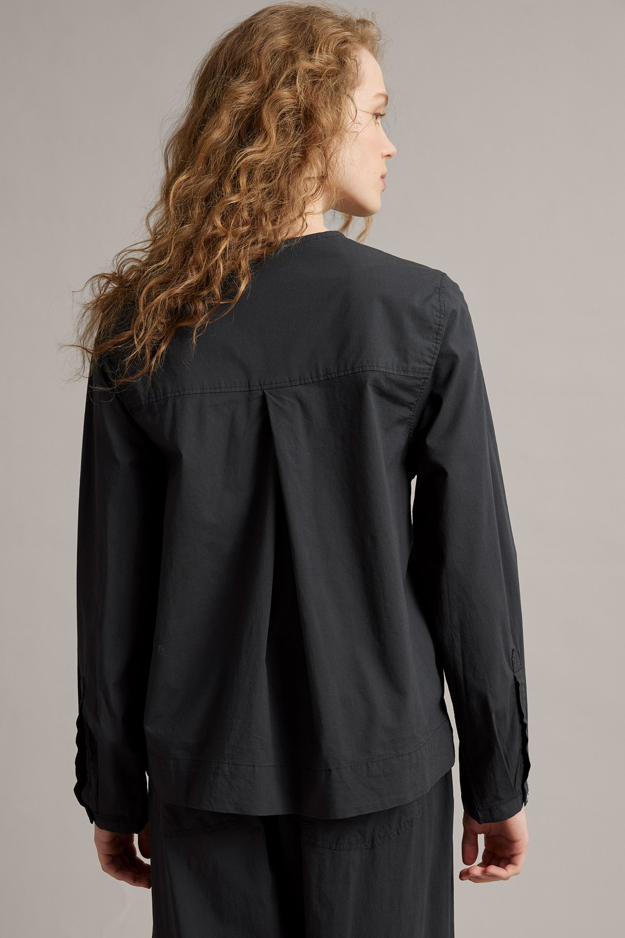 Naisen takasivukuva mustassa, löysässä puuvillapopliinipuserossa, jossa on pitkät hihat ja laskokset takana.