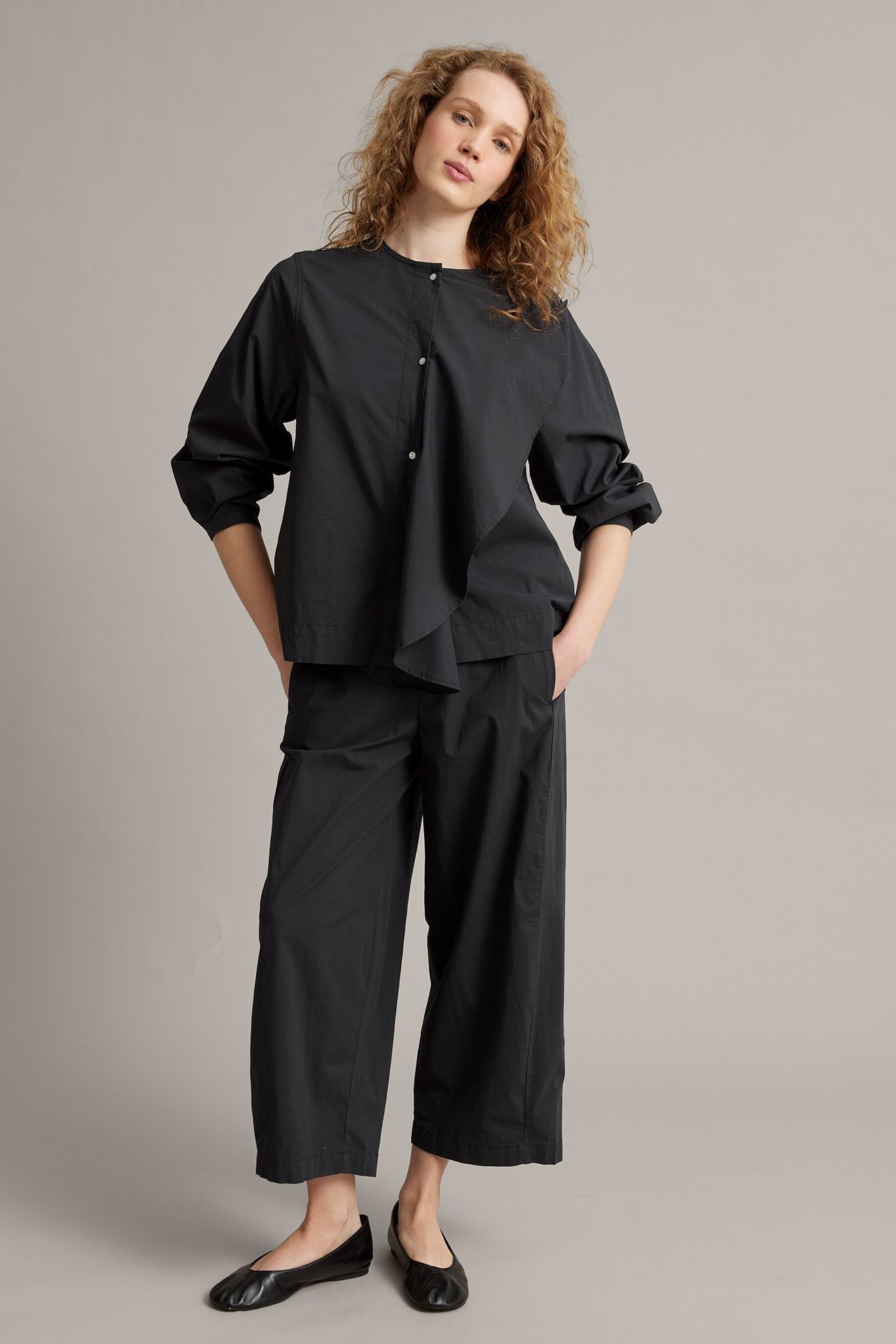 Frontbilde av kvinne i svart poplinbluse med tre knapper, svarte cropped bukser og svarte ballerinasko.