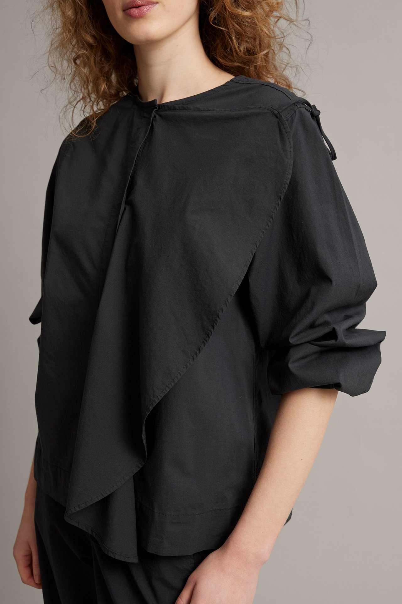 Edestä, nainen yllään löysä musta popliinipusero, jossa suuri drapeerattu röyhelö ja olkainnauhat.