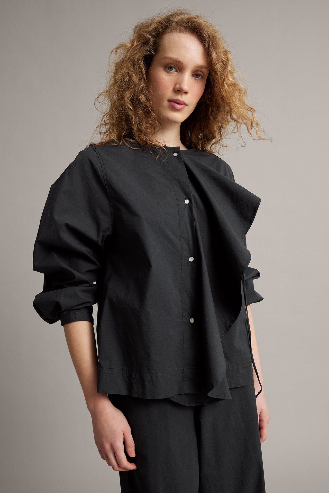 Widok z przodu kobiety w czarnej bluzce popelinowej z odpinaną falbanką, stylizowanej z czarnymi spodniami.