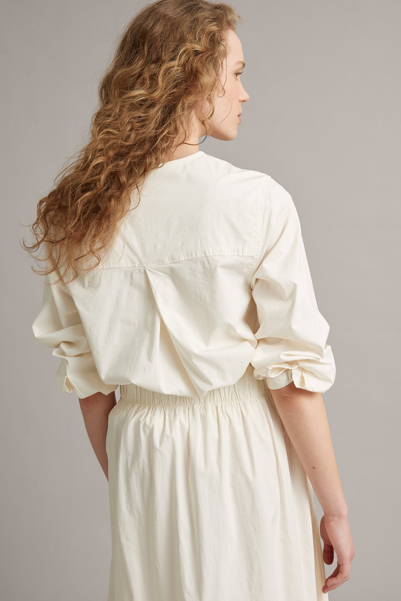 Naisen sivulta takaa: valkoinen popliinipusero käärityillä hihoilla ja yhteensopiva hame.