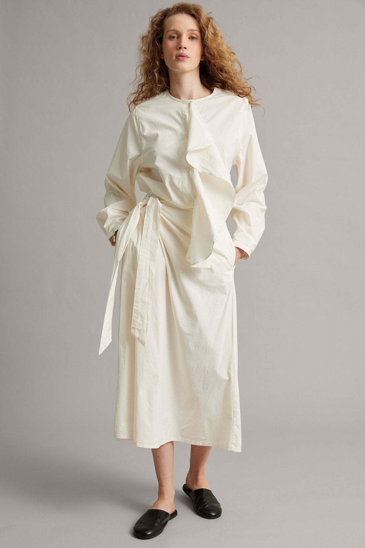 Forfra: kvinne i lang hvit poplinbluse med rysje, knytebånd i midjen og svarte slip-on sko.