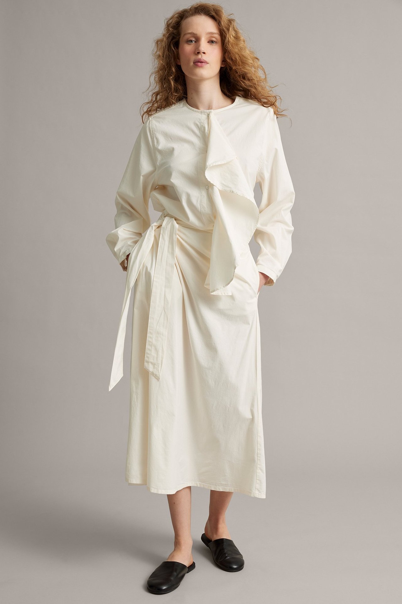 Forfra: kvinne i lang hvit poplinbluse med rysje, knytebånd i midjen og svarte slip-on sko.