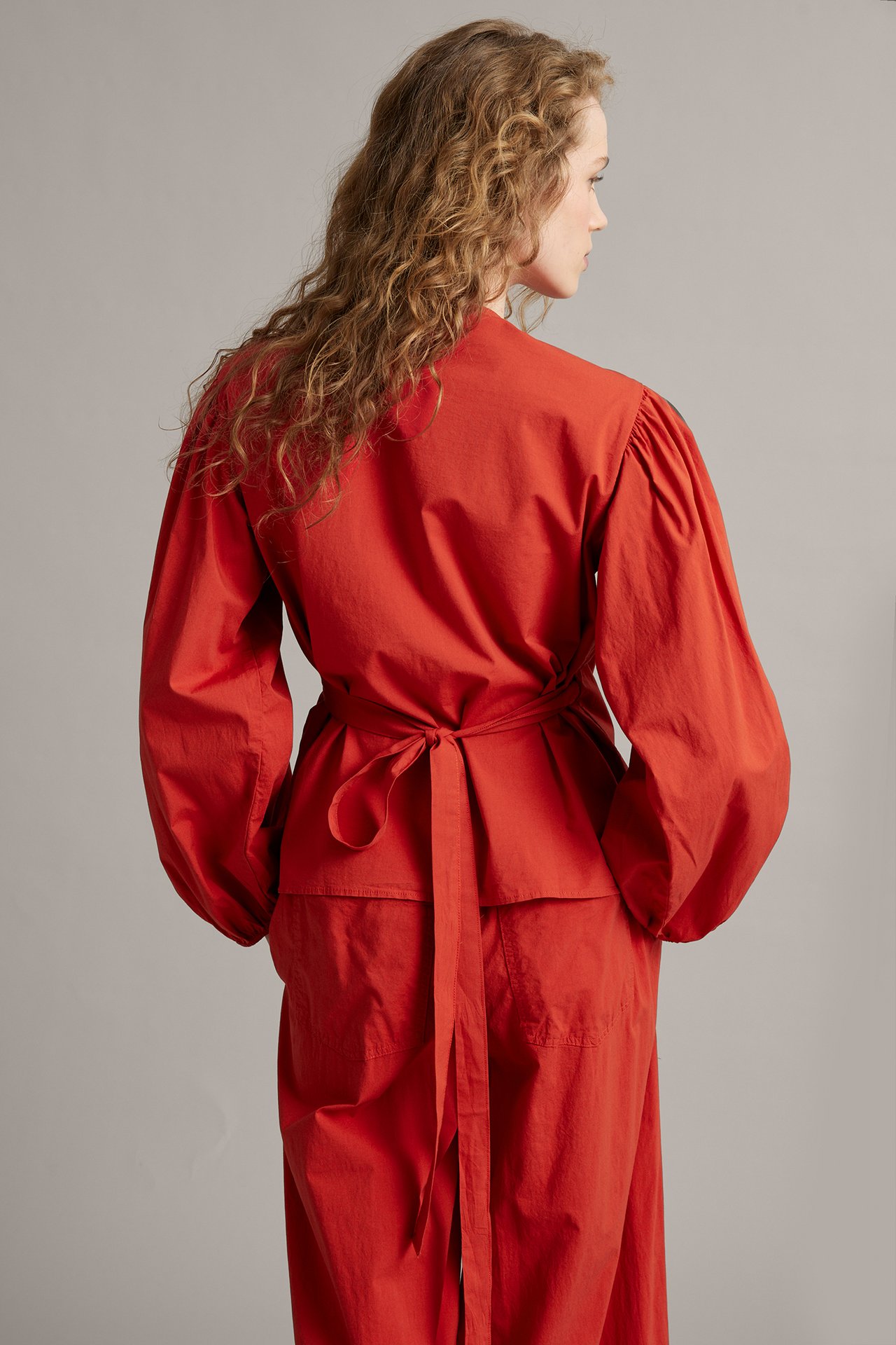 Takaa: nainen punaisessa kietaisublousessa volyymihihoilla, sidottu, ja yhteensopivat leveät housut.