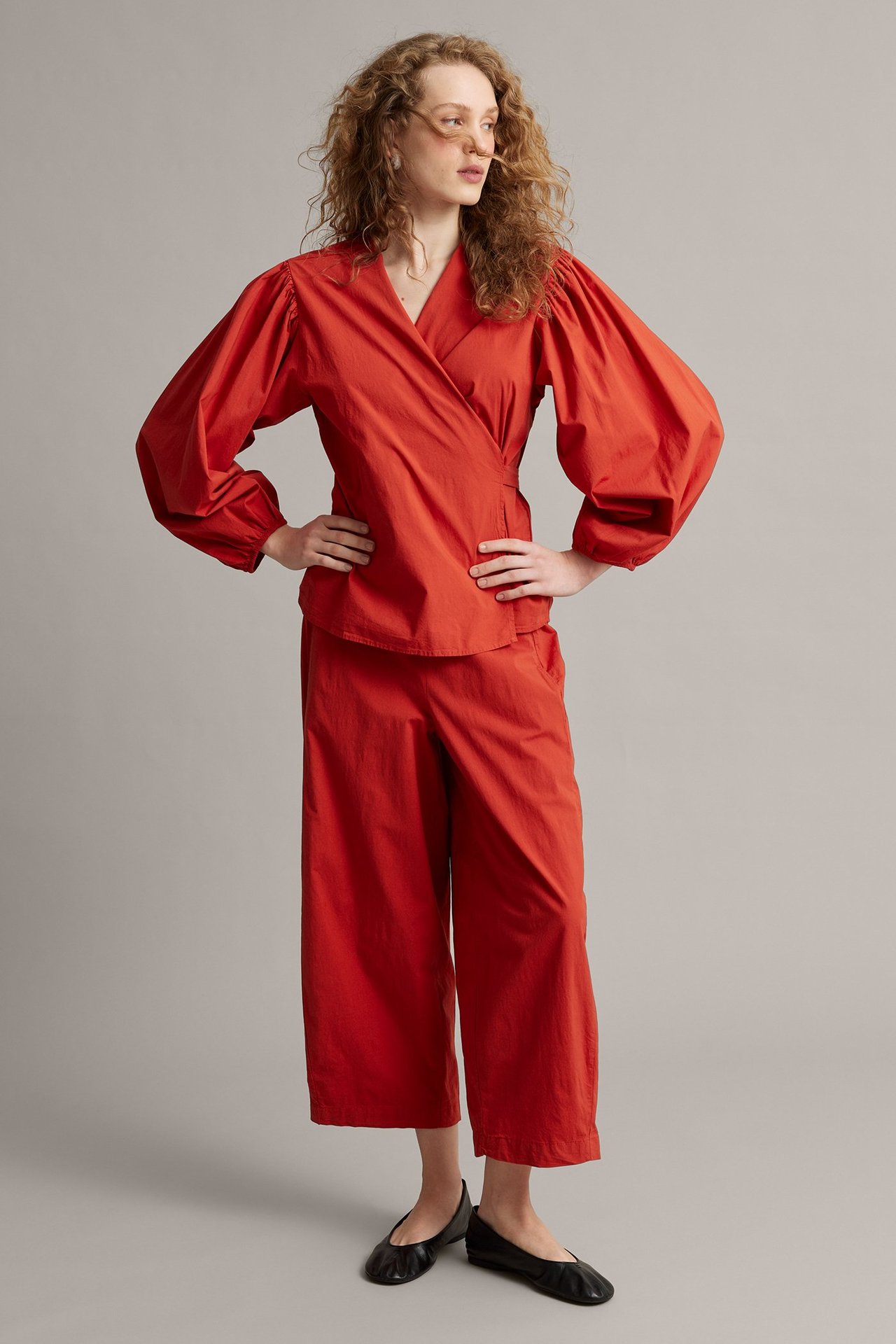 Forfra: Kvinne i rød omslagsbluse i poplin med ballongermer, matchende croppete bukser, svarte ballerinasko.