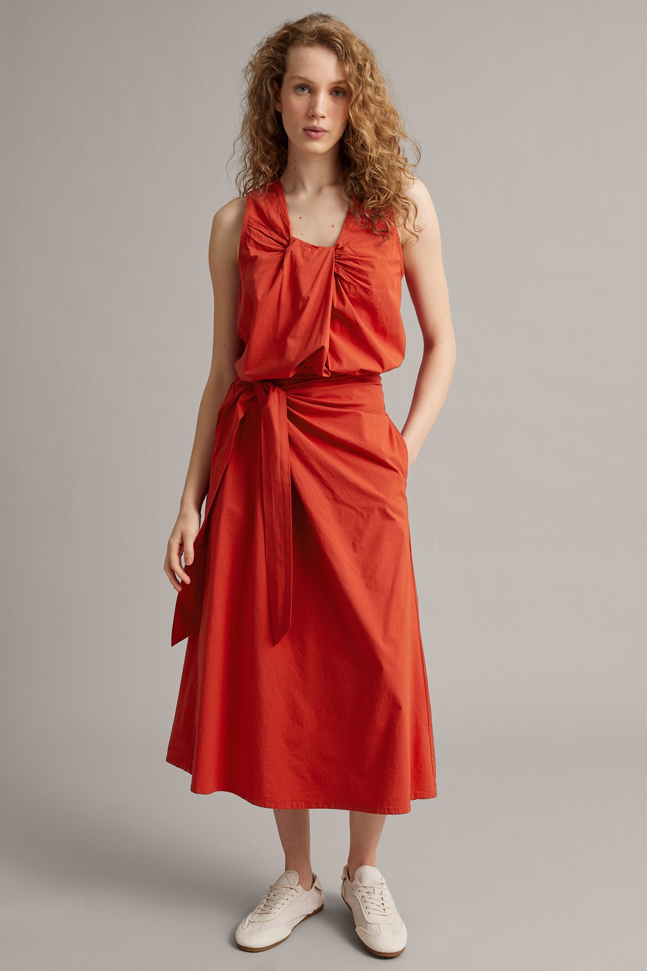 Framifrån en kvinna i röd omlottkjol med knyt i poplin, matchande ärmlös topp och ljusa sneakers.