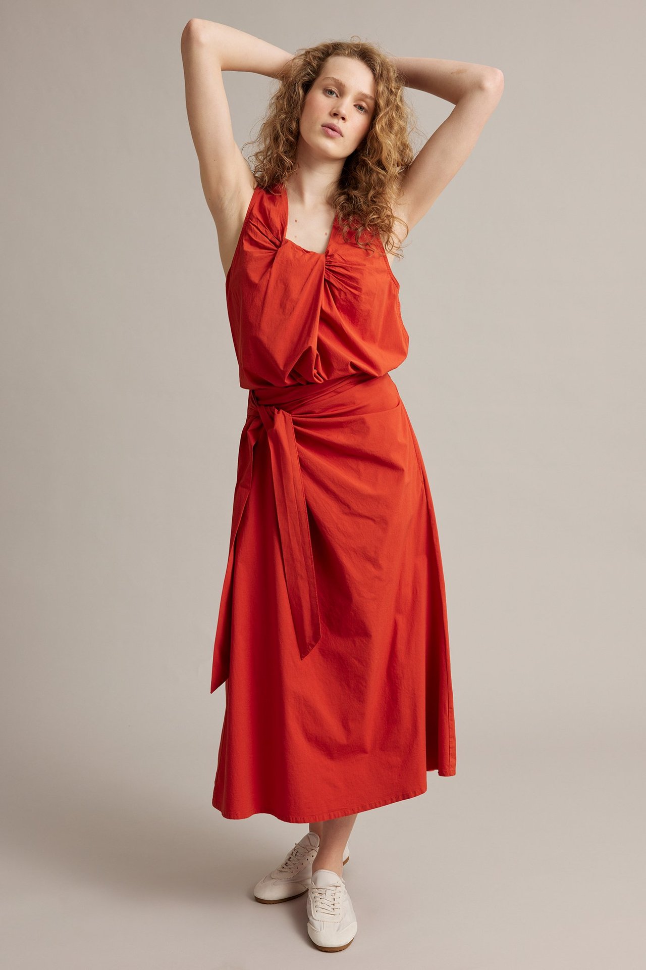 Widok z przodu: kobieta w czerwonej spódnicy kopertowej z popeliny z wiązaniem, dopasowanym czerwonym topie i białych sneakersach.