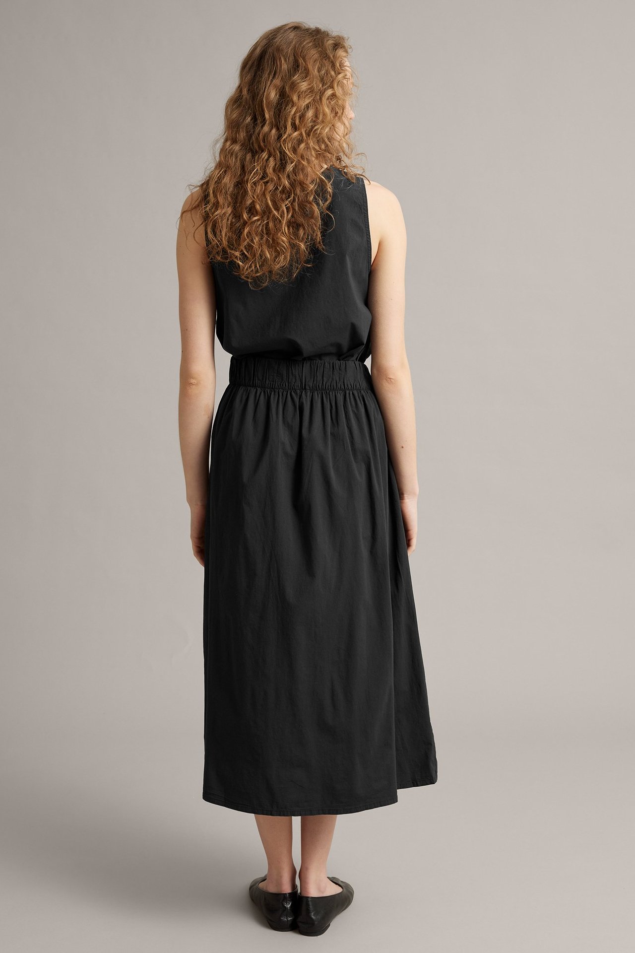 Nainen takaa, yllään musta A-linjainen kietauluhame joustavalla takaosalla, musta hihaton toppi ja mustat ballerinat.