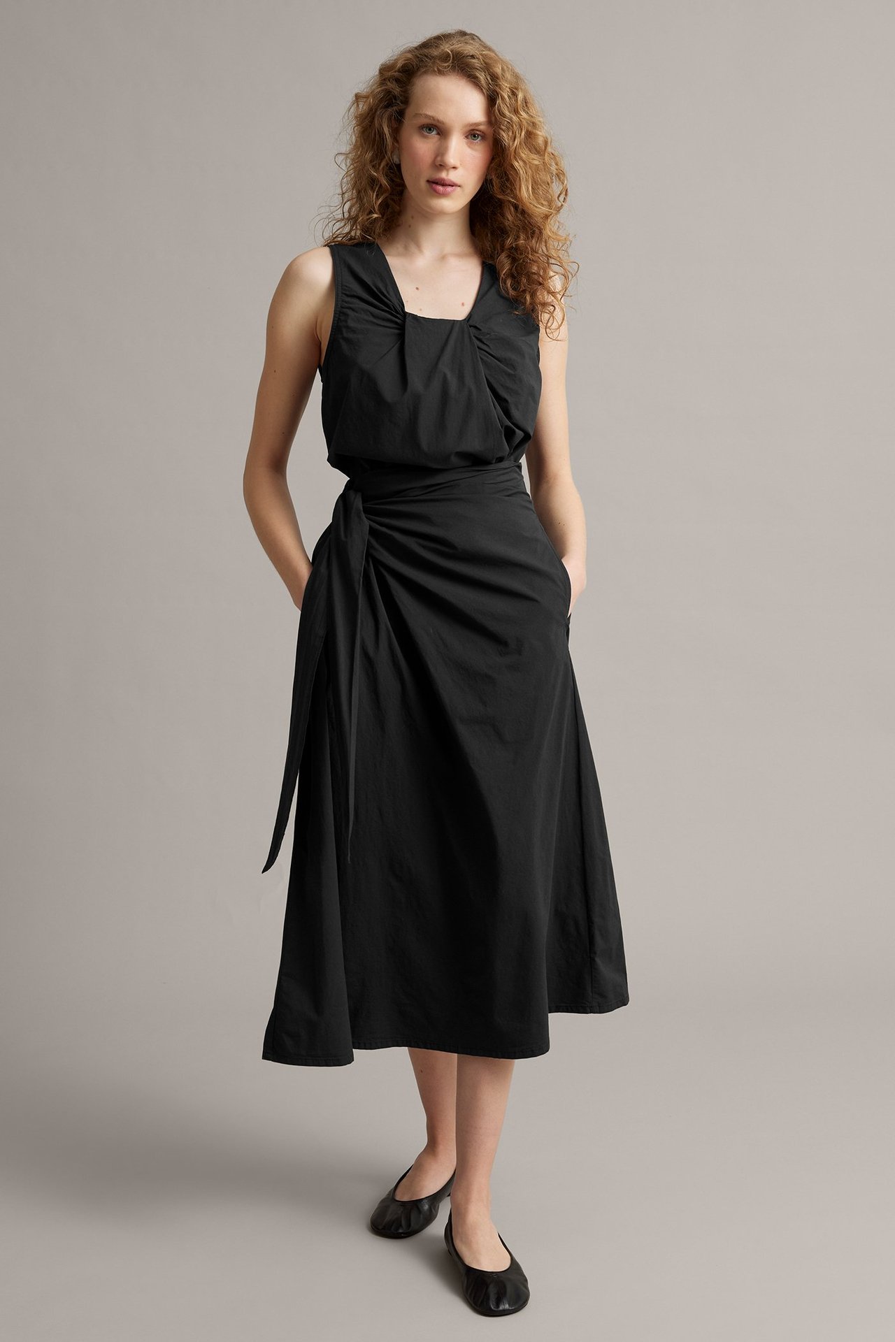 Forfra, kvinne i svart poplin omslagsskjørt med knyting i siden, matchende topp og svarte ballerinasko.