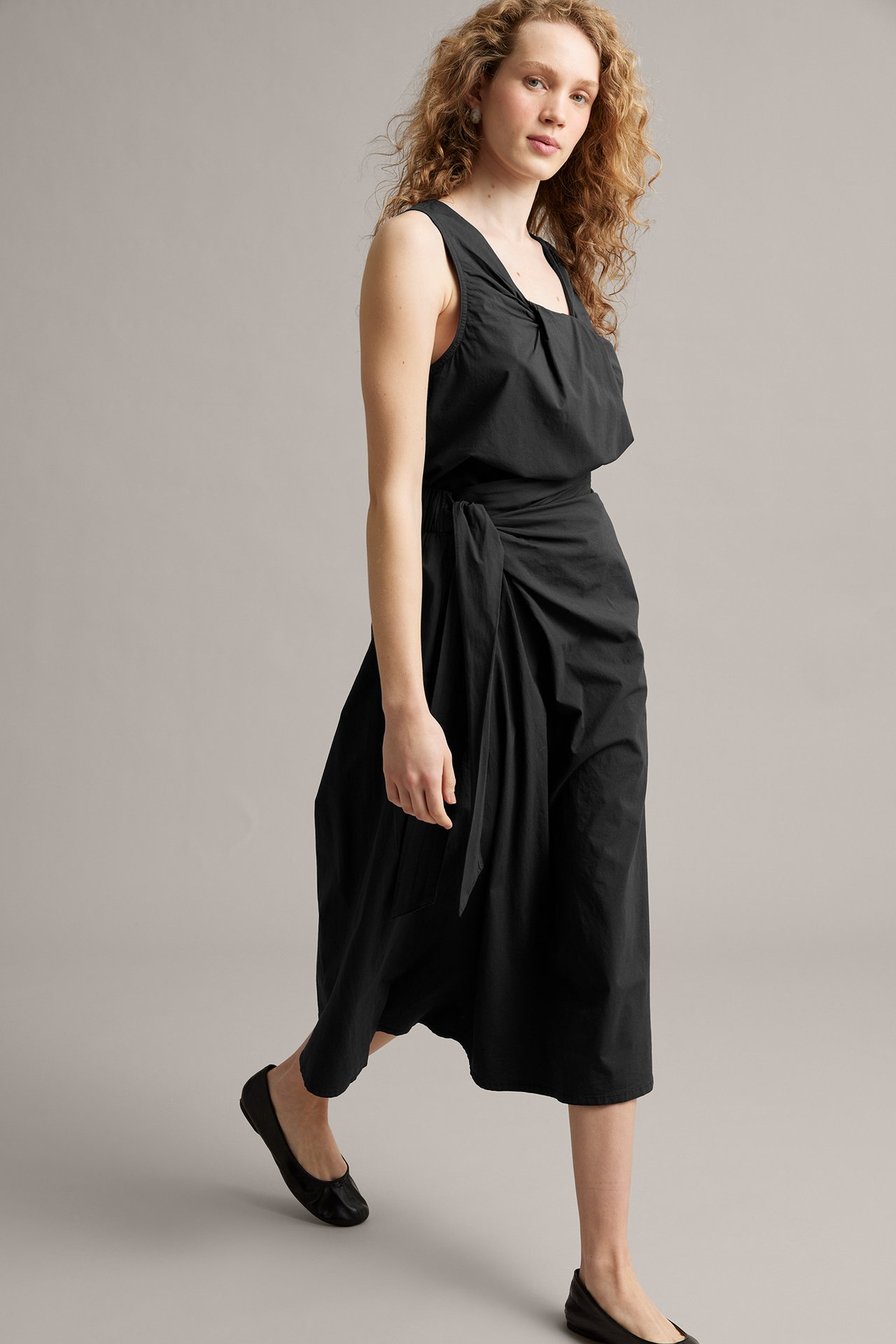 Naisen sivukuva mustassa popliinihameessa, jossa solmiminen, mustassa hihattoman topin kanssa ja ballerinoissa.