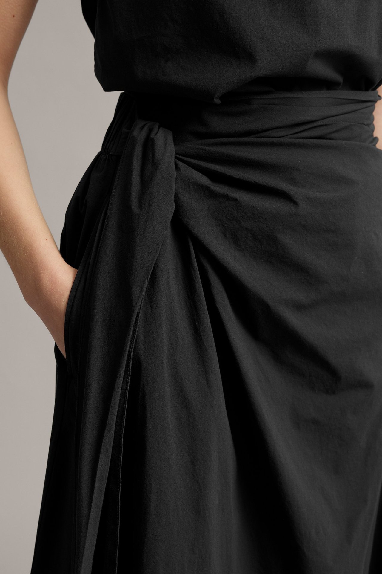 Lähikuva edestä naisesta, jolla on musta popliininen kietauluhame sivusolmiolla ja musta toppi.