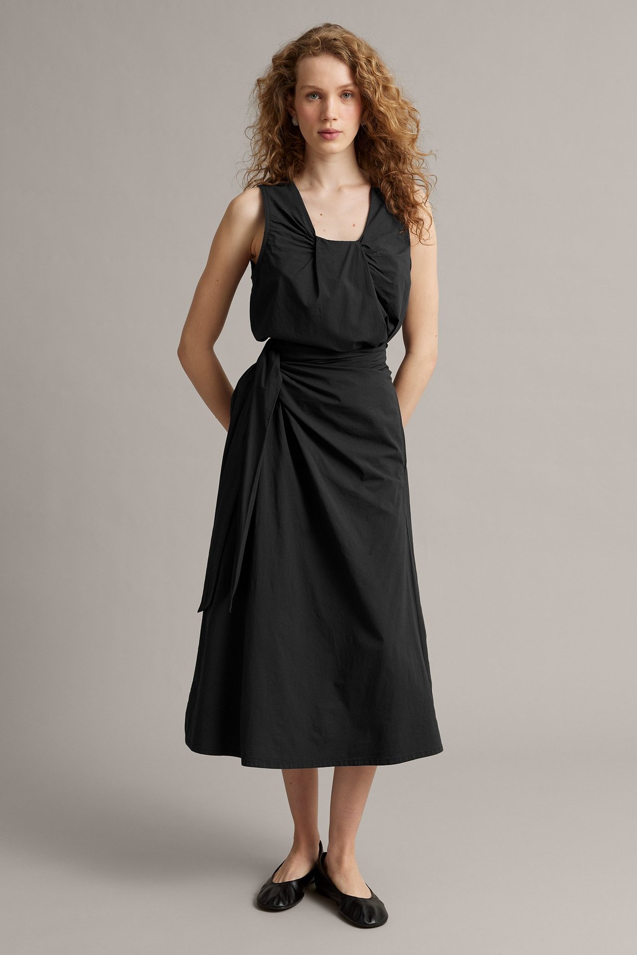 Kobieta w czarnej popelinowej spódnicy kopertowej z wiązaniem, topie bez rękawów i balerinach.
