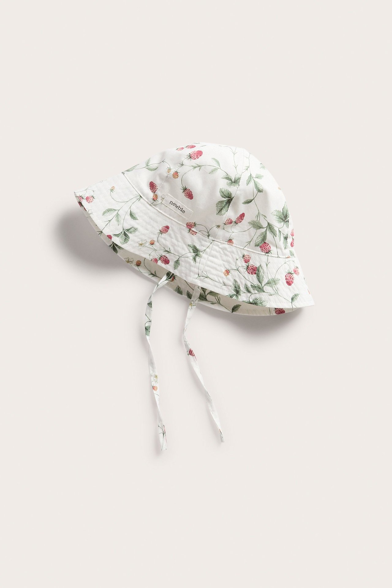 Yläkulmasta kuvattu luonnonvalkoinen puuvillainen aurinkohattu mansikkakuviolla ja sidontanauhoilla.