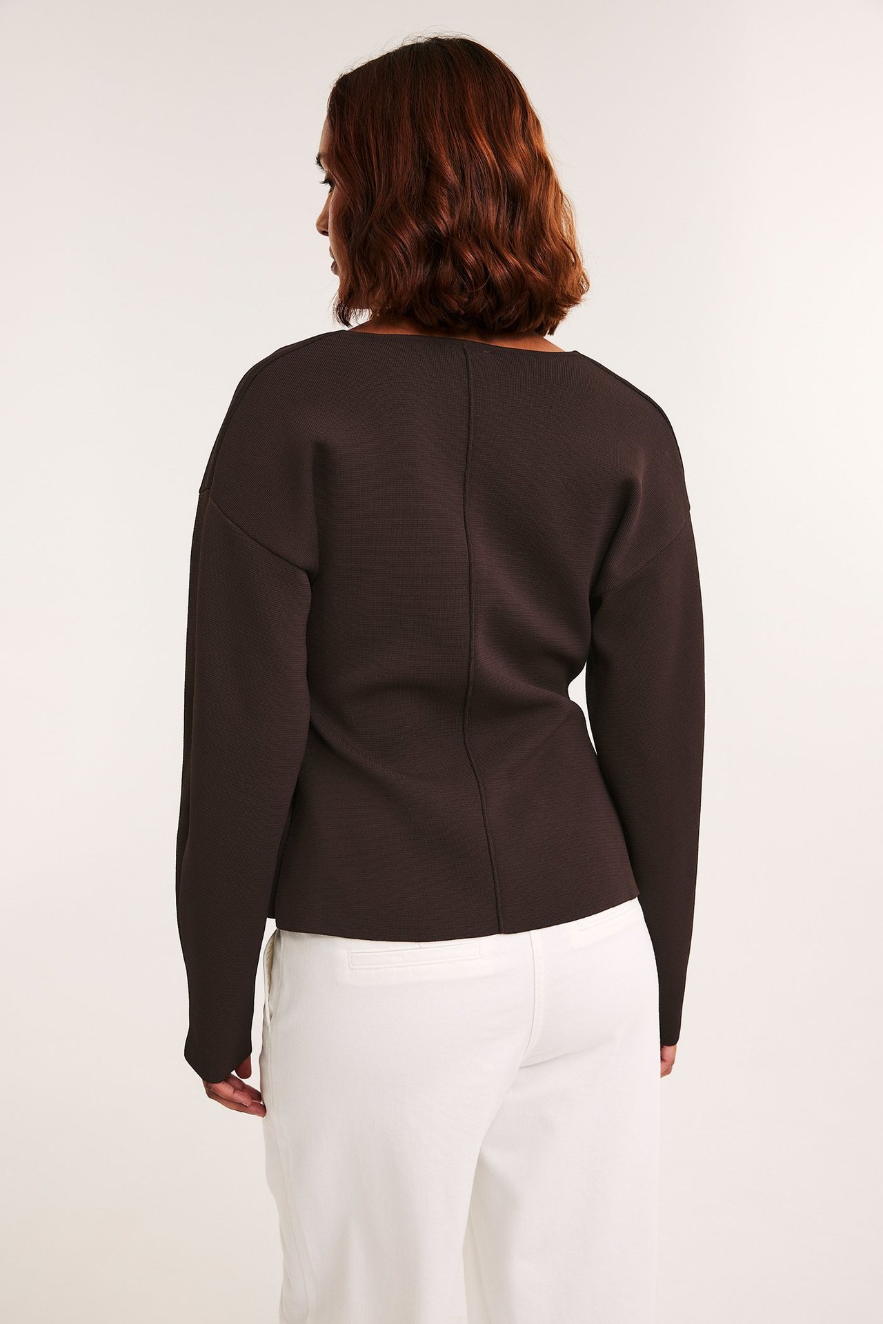Ryggvy av en kvinna i en mörkbrun figursydd långärmad topp och vita byxor.